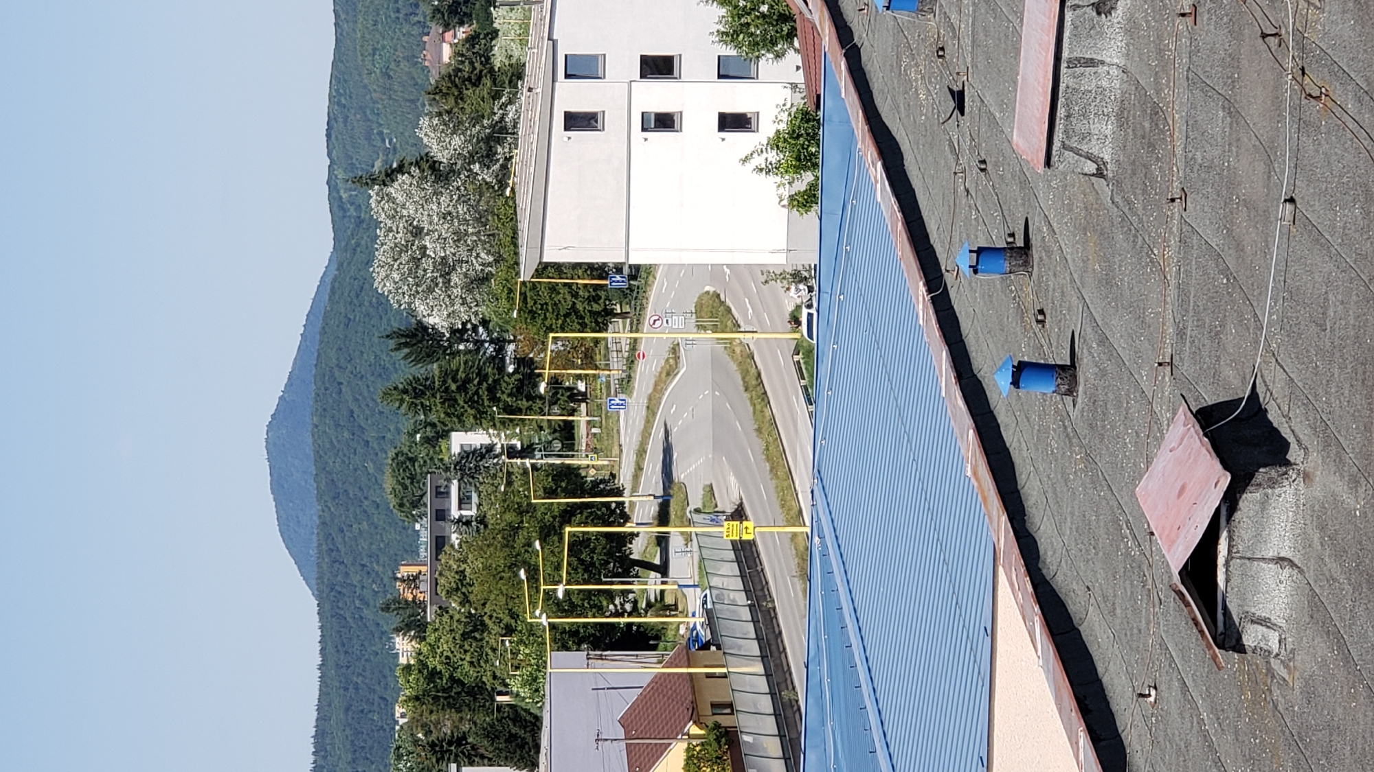 This image depicts a split view of a city scene. The upper portion captures a panoramic view of a residential area with a road curving through it, bordered by trees and buildings. In the background, a large, forested hill dominates the skyline under a clear blue sky. The lower portion focuses on the rooftop of a building, showing several ventilation pipes, skylights with red, angular covers, and electrical wiring. The scene appears to be in Prešov, Slovakia, with the prominent hill in the background likely being Zlatá baňa. The image captures a daytime scene, with bright sunlight casting shadows. There are no visible people in the image.
