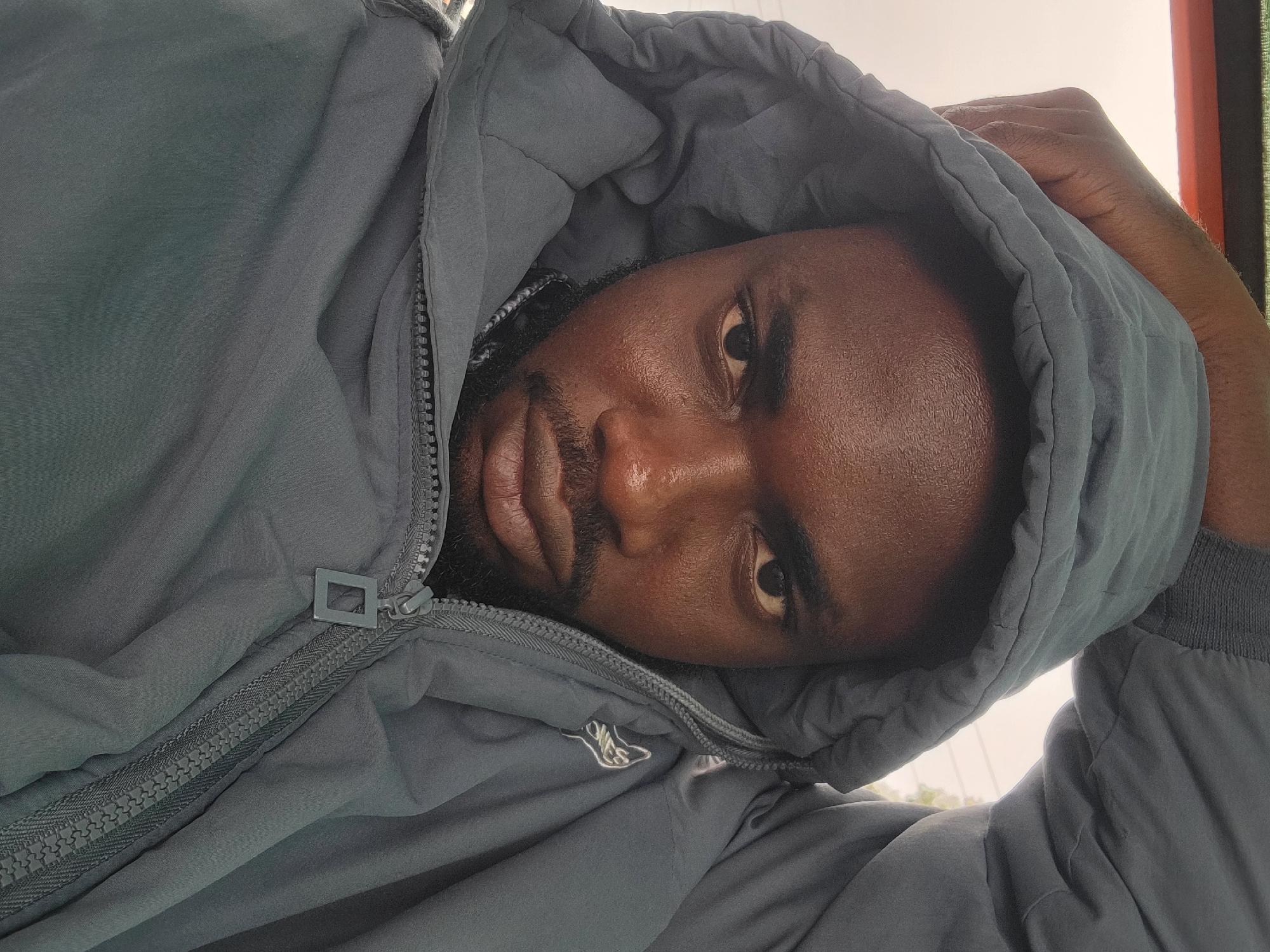 The image shows a close-up selfie of a man wearing a gray hooded jacket. He has dark skin, a beard, and is looking directly at the camera with a serious expression. His left arm is raised, with his hand resting on the hood of the jacket, pulling it over his head. The jacket is zipped up partially, revealing a glimpse of a patterned fabric underneath. The background is blurred and appears to be an outdoor setting with a light-colored sky and some faint green foliage, suggesting it might be an urban or semi-urban environment. No specific location in Gura Top, Nigeria, can be identified from the image.