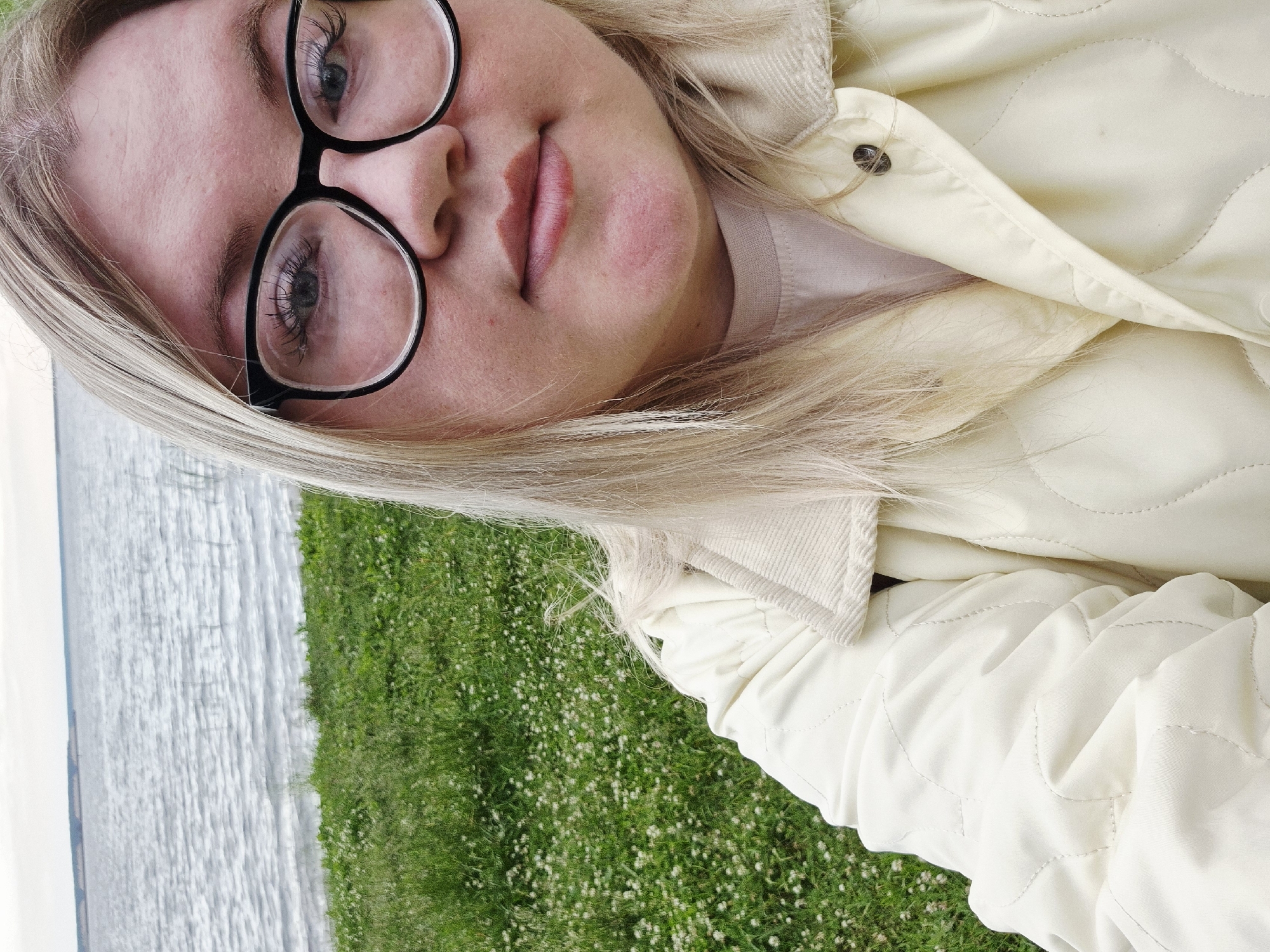 In Chaikovsky, Russia, a young woman with blonde hair and glasses is taking a selfie by a body of water. She is wearing a light-colored jacket over a pink t-shirt. The background features a grassy area with small white flowers and a calm body of water that stretches to the horizon, where a faint landmass is visible. The sky is overcast.