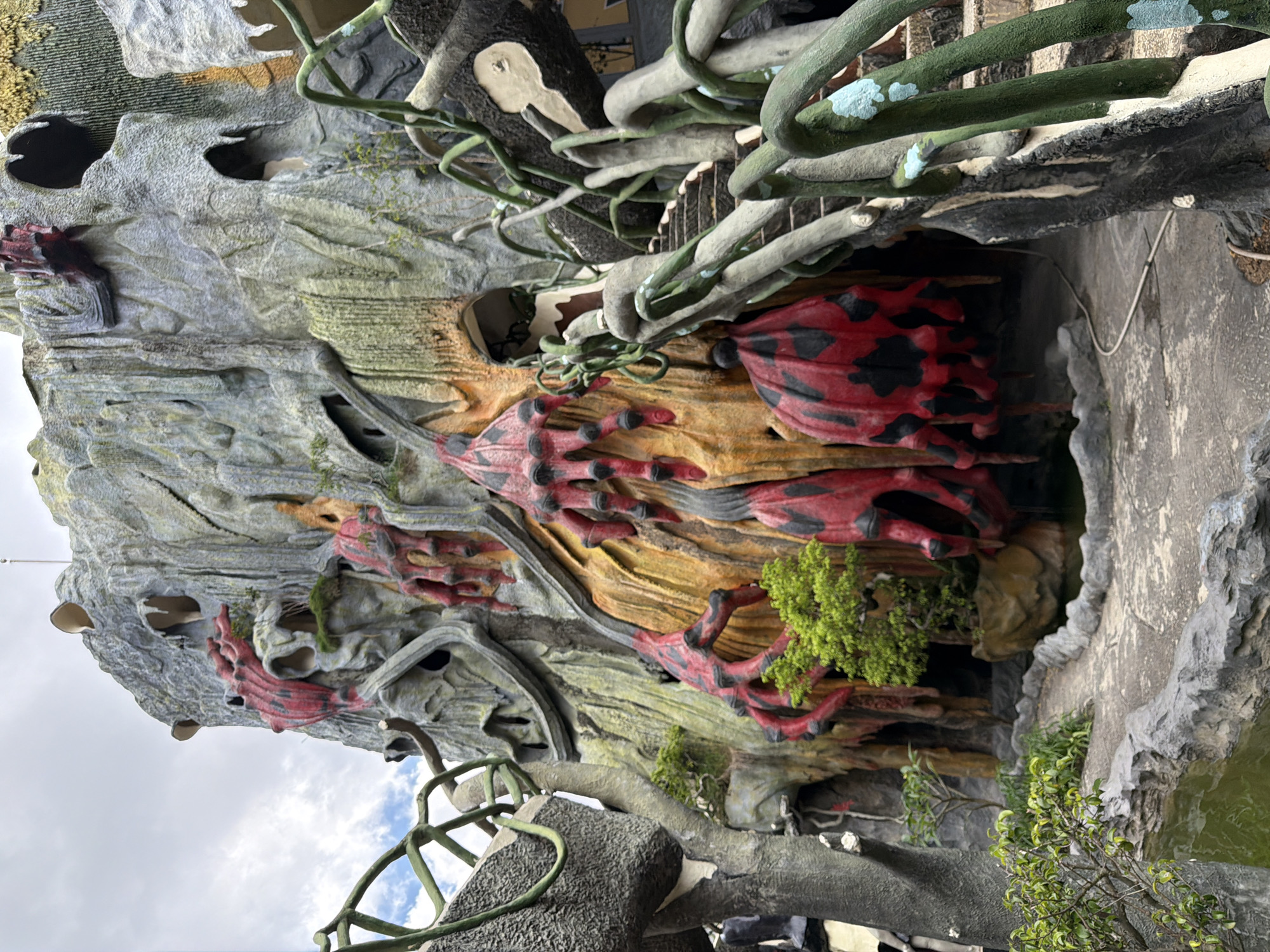 The image depicts a section of the exterior of the Hang Nga Guesthouse, also known as the "Crazy House," in Da Lat, Vietnam. The architecture is organic and surreal, resembling a giant, gnarled tree or a natural cave formation. The dominant colors are gray and moss green, with accents of red and yellow. Numerous bulbous, red, hand-like appendages with black spots emerge from the textured gray and yellow walls. Twisted, vine-like structures, painted in green and gray with occasional light blue accents, form railings and decorative elements, some of which frame a stone staircase leading upwards. Green foliage, including small trees and bushes, is interspersed throughout the scene, growing from crevices and along pathways. A small pool of water is visible in the foreground, bordered by rough, gray rock formations. The sky above is overcast with clouds. There are no people visible in the frame. The overall impression is one of a fantastical, somewhat unsettling, and otherworldly environment.