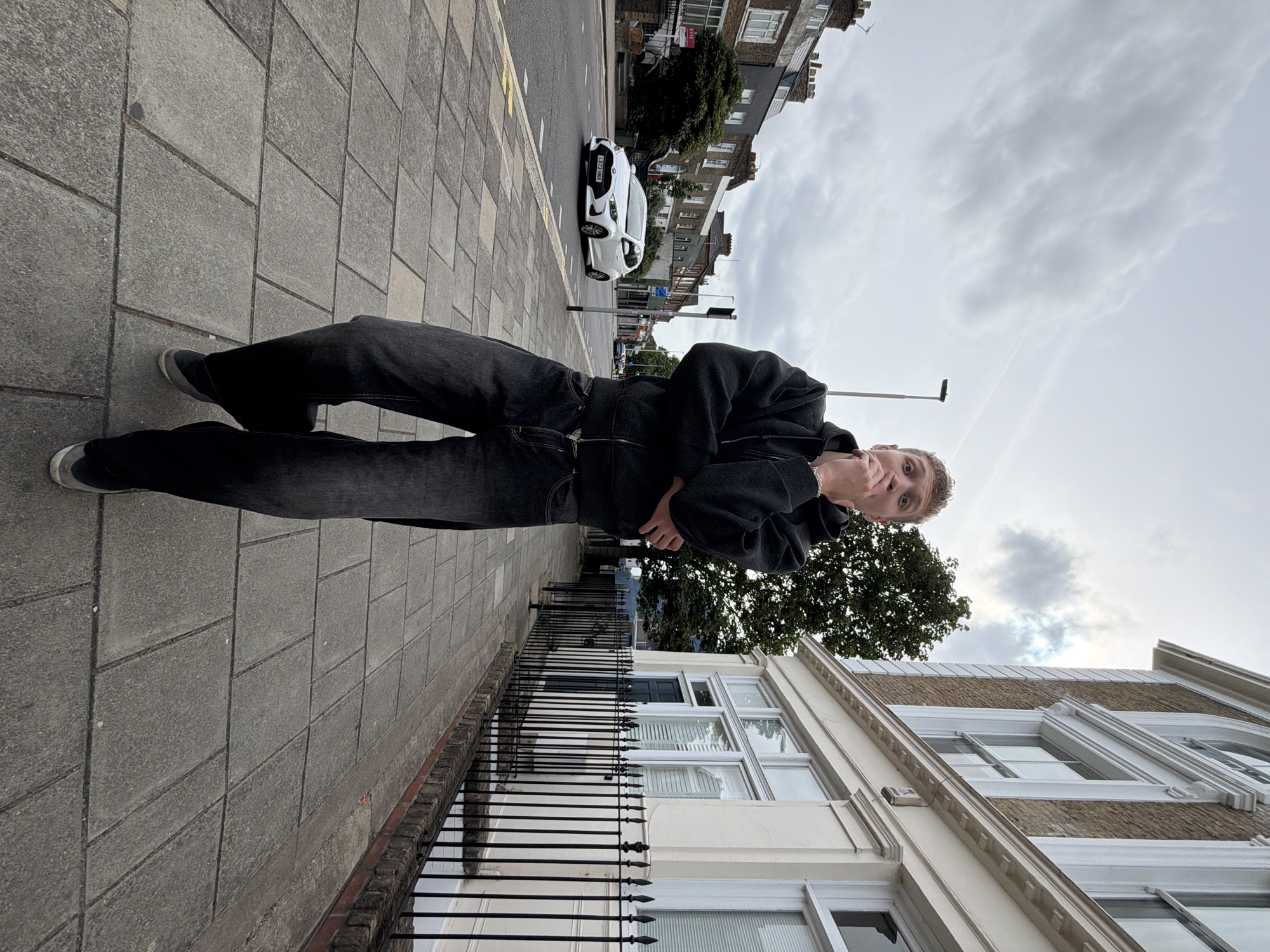 This is a full-length, low-angle outdoor photograph of a young adult male standing on a paved sidewalk on an overcast day. The subject, with short blonde curly hair and light skin, is positioned centrally, facing the camera. He wears a dark grey zip-up hoodie, faded black wide-leg jeans, and dark grey sneakers with white soles. His right hand is placed over his mouth, and his left arm is folded across his chest.
The sidewalk, constructed from grey rectangular paving slabs, runs parallel to a two-lane asphalt road. On the road to the subject's left, a white Toyota Yaris, bearing the UK license plate 'LB73 HNX', is visible moving away from the camera. Across the road, a row of multi-story terraced houses with brick facades and tiled roofs extends into the background; one house displays a 'KFH' real estate sign. To the subject's right, a substantial multi-story building features light-colored brickwork, large white-trimmed sash windows, some with arched tops, and decorative architectural details. A black wrought-iron fence separates this building from the sidewalk. A large, leafy green tree is visible behind the fence and building. The sky is entirely covered by grey clouds, and a faint airplane contrail is visible in the upper sky.
The architecture, the presence of the UK vehicle registration plate 'LB73 HNX' (where 'LB' signifies registration in the Wimbledon/Kingston area), and the 'KFH' real estate agent sign (a London-based firm) collectively identify the location as an urban street in London, United Kingdom, specifically within South West London.