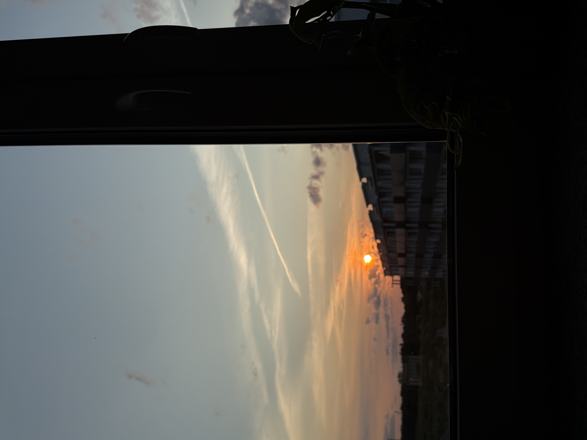 The image captures a sunset viewed through a window in Gorzow, Poland. The sun, a bright orange orb, is low on the horizon, casting a warm glow across the sky. Wisps of clouds streak across the pale blue and orange sky, with a prominent contrail visible. In the middle ground, a silhouetted apartment building stretches horizontally, its details softened by the fading light. The foreground is dominated by the dark silhouette of the window frame and a houseplant, obscuring the lower portion of the scene. The overall impression is one of a peaceful, urban twilight.