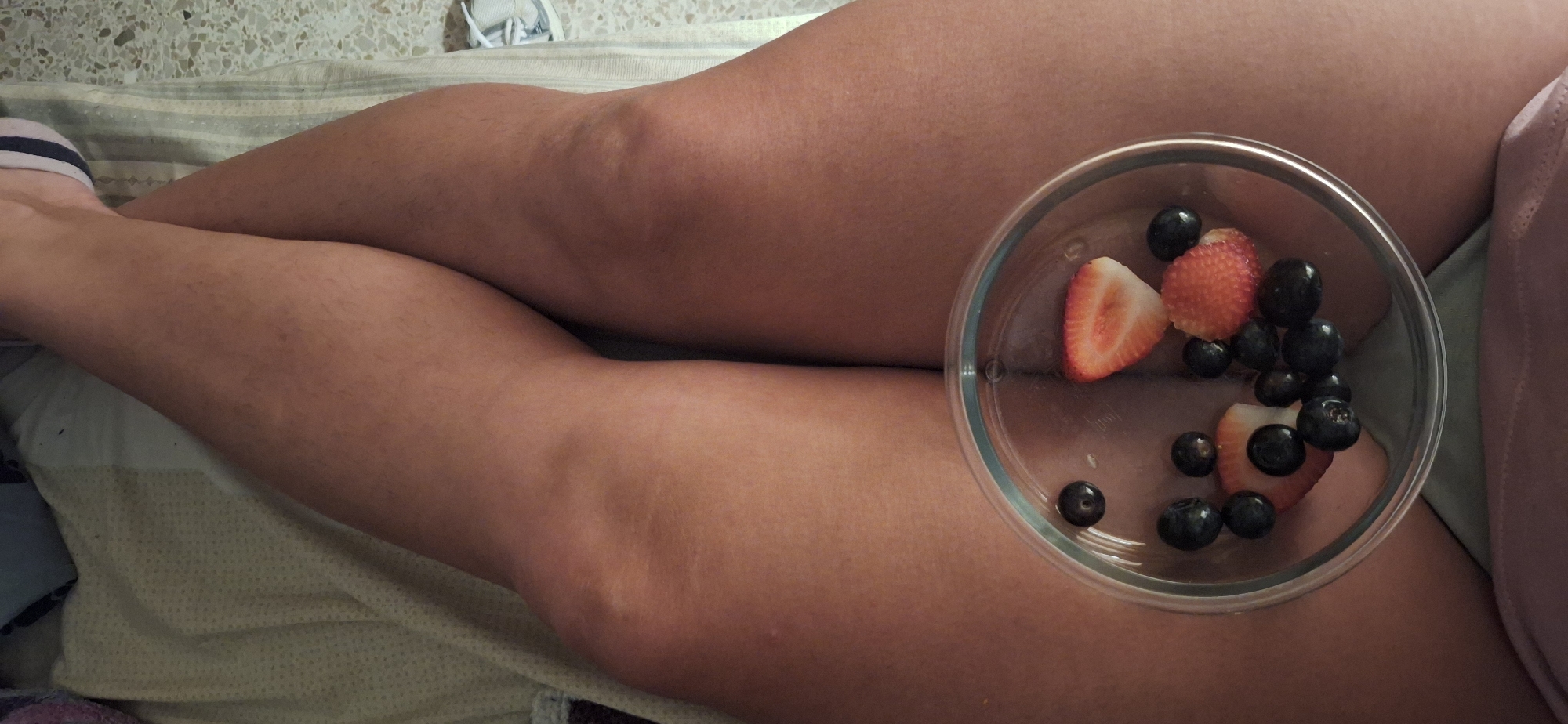 A high-angle, close-up shot captures a person's legs, likely bare, resting in a relaxed position. The legs are seen from the thighs down to the calves, with a light-colored blanket or sheet partially visible beneath them. A clear glass bowl containing a mix of strawberries and blueberries rests on one of the upper legs. The strawberries are cut into halves, and there are several dark blueberries scattered within the bowl. A small portion of a foot wearing a white sock is visible in the bottom-left corner of the frame. The background shows a light-colored, textured surface, possibly a wall or floor. The scene depicts a moment of leisure, possibly enjoying a healthy snack while relaxing indoors in Santa Cruz De Tenerife, Spain. No visible text is present in the image.