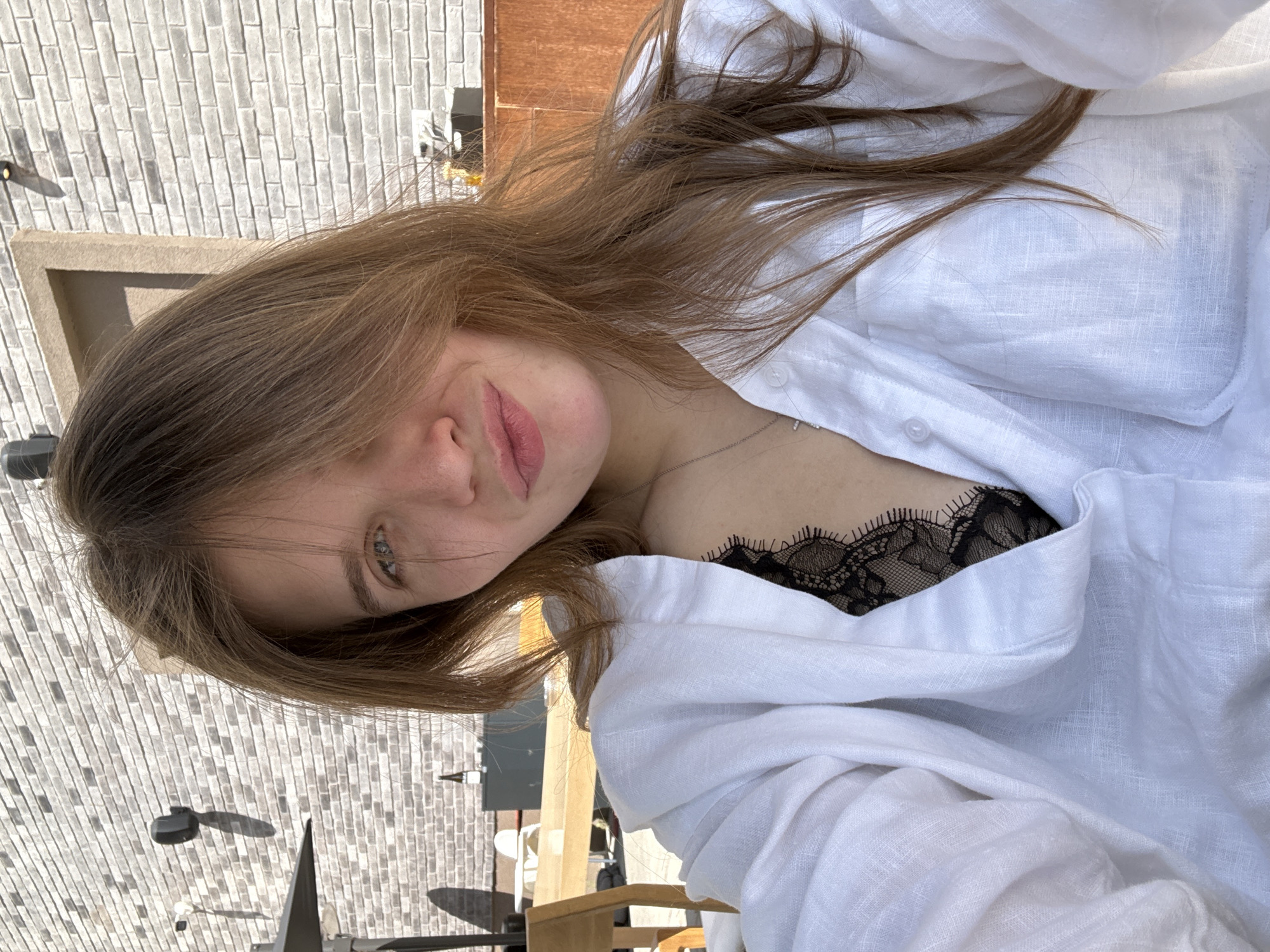 A young woman poses for a selfie in an outdoor setting in Belgrade, Serbia. The background shows a brick wall, a wooden chair, and a portion of an awning. The woman is wearing a white linen shirt, slightly unbuttoned to reveal a black lace garment underneath. She has light brown hair, and her face is partially obscured by her hair. Her expression appears relaxed.