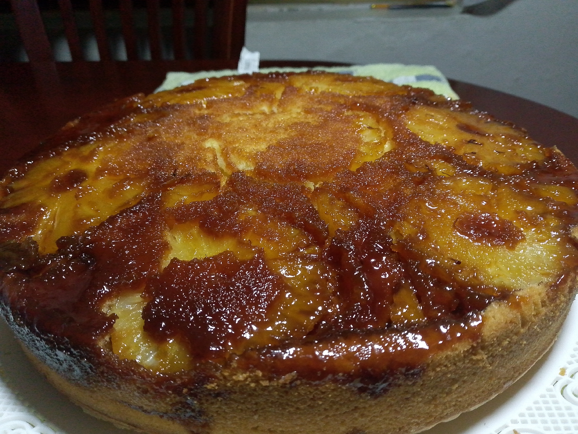 A close-up shot of a freshly baked pineapple upside-down cake on a white doily. The cake is round and sits on a dark, possibly wooden, table. A partially visible dark wood chair is in the background, along with a light-colored surface. The cake itself features caramelized pineapple pieces embedded in a rich, brown, sugary topping. The cake portion is golden brown and slightly textured. The image appears to be taken indoors with soft, diffused lighting. There is no information to confirm this photo was taken in Tinaquillo, Venezuela.