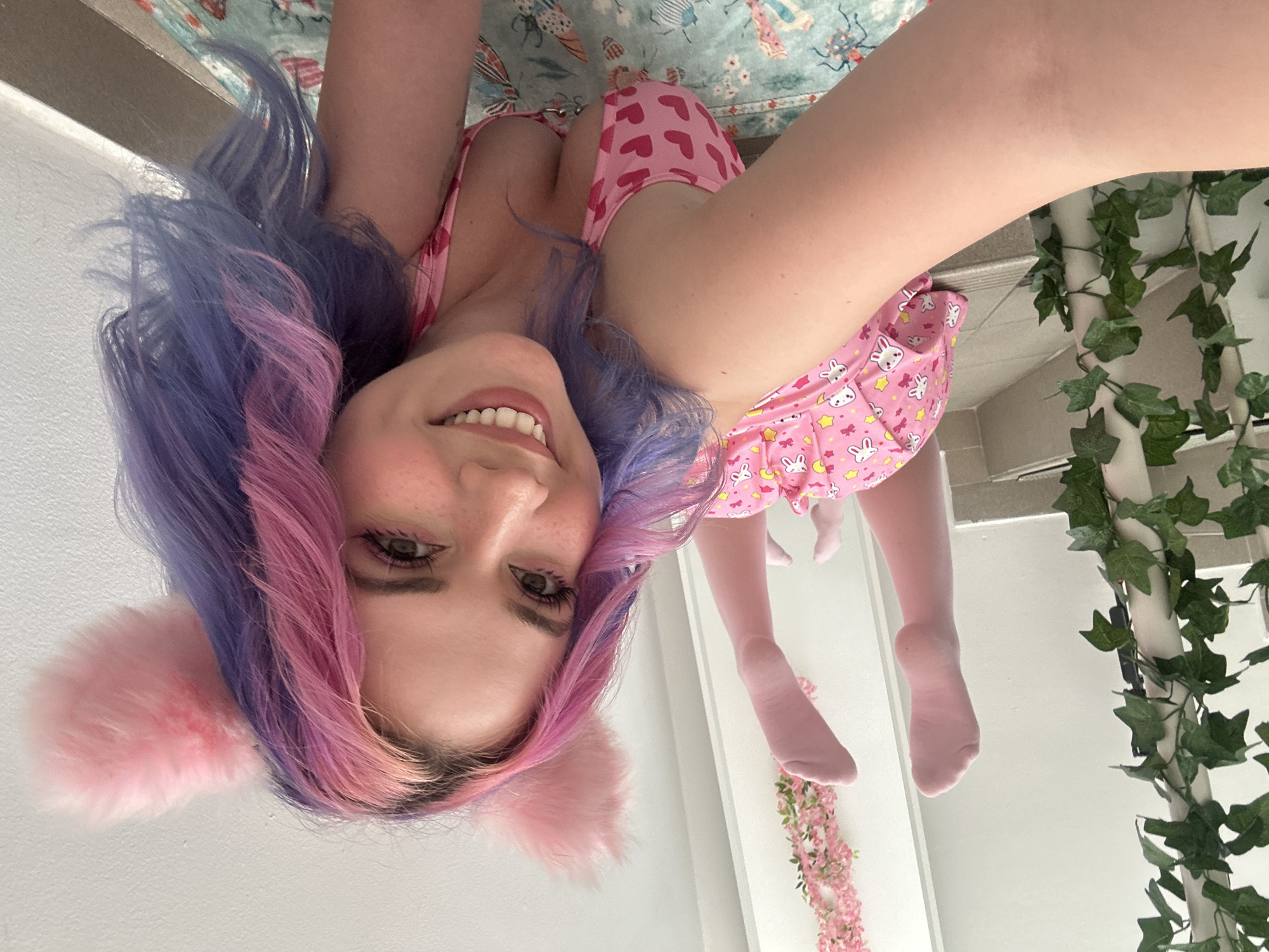 A young woman with colorful hair (purple and pink) and cat ears is positioned upside down in what appears to be a room, possibly in the City of Zagreb, Croatia. She is wearing a pink outfit, including what looks like a romper with a pattern of rabbits and stars, and pink tights. The background includes a rug with a pattern of insects and other designs, and a wall decorated with artificial green vines and a pink flower arrangement. The walls are white, and there is what appears to be a door visible in the background.