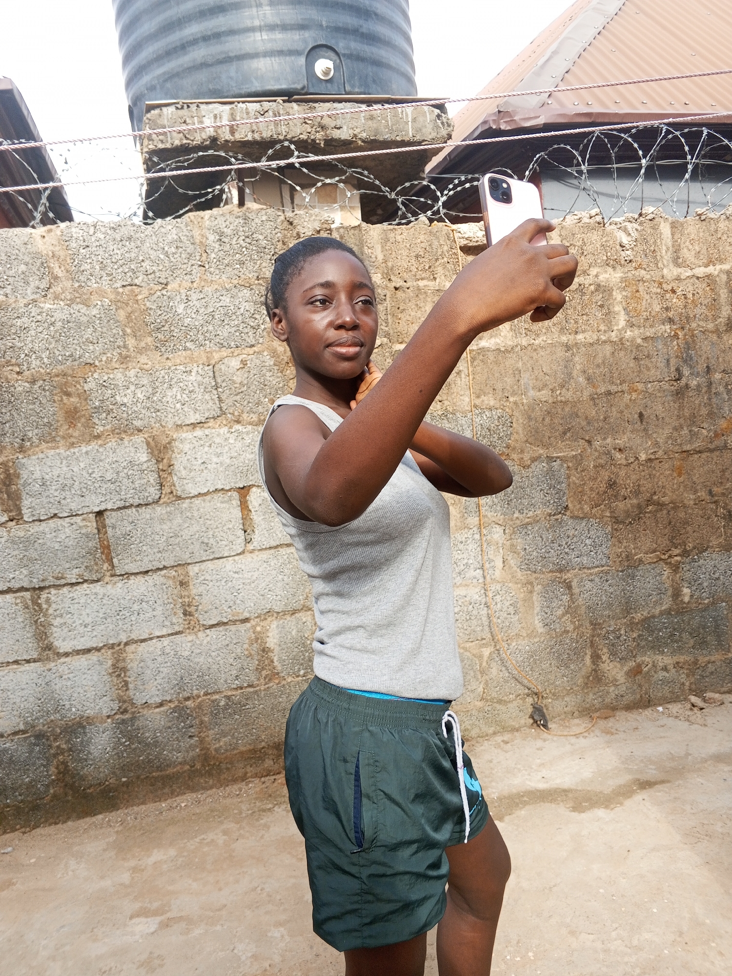 A young woman stands outside, holding a smartphone as if taking a selfie. She wears a grey tank top and dark green shorts with a blue and white logo. Behind her is a cinder block wall topped with barbed wire, and above that, a water tank and part of a metal roof. The ground is dirt and concrete. The location is likely in a residential area of Abuja, Nigeria, indicated by the construction style and infrastructure elements.