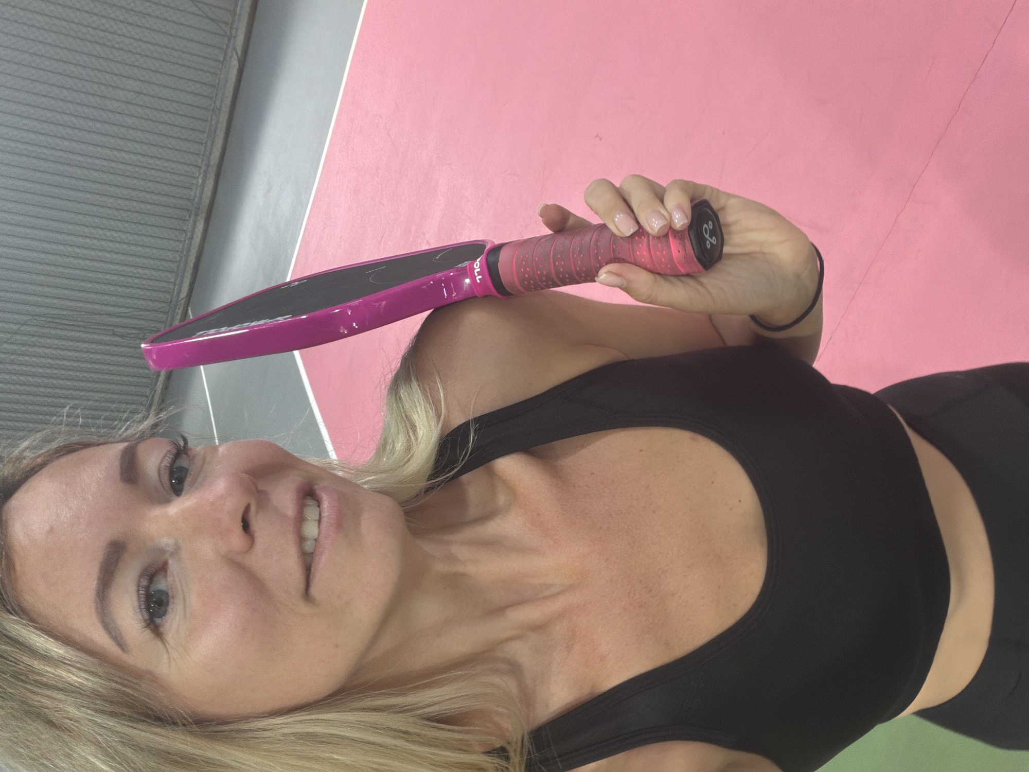 A blonde woman in a black sports bra holds a pink paddle and stands on a pink court. The paddle has a black grip with pink accents and is branded with "TYE". The woman has blue eyes and a slight smile, and her fingernails are painted a light pink. The background features a grey wall with vertical lines, likely a net or enclosure. The court surface is divided into pink and green sections. The lighting suggests an indoor setting.