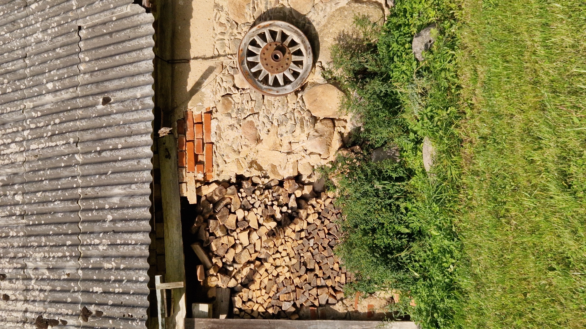 A weathered, corrugated asbestos roof covers the top of a rustic stone and brick building. Below the roofline, a section of stacked firewood is visible, piled neatly against the rough stone wall. To the right of the firewood, a vintage wooden wagon wheel with a rusted metal hub is mounted on the stone wall, its spokes radiating outwards. Dense green foliage and grass grow at the base of the building, partially obscuring some scattered rocks. The scene appears to be outdoors in a rural or semi-rural setting, possibly in Tāši, Latvia, given the architectural style and the presence of natural elements.