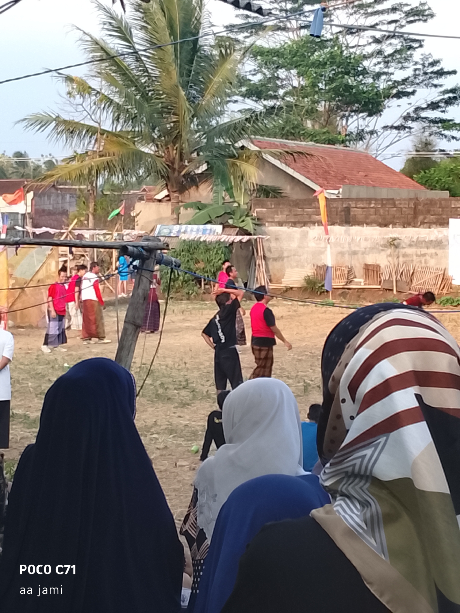 The image depicts an outdoor scene in Tasikmalaya, Indonesia, likely a community event or gathering. In the foreground, three individuals wearing headscarves are partially visible from behind, suggesting they are spectators. The person on the left wears a dark blue hijab, the middle person a white hijab, and the person on the right a patterned hijab with brown, white, and beige stripes.
Further into the scene, a group of people are engaged in an activity, possibly a game or a performance, on a dirt ground. Several young men and boys are present. Some are wearing red shirts or vests, and many are wearing sarongs or long patterned pants. One individual in the middle ground is seen running or in motion. A wooden pole with wires is prominent in the middle ground, acting as a visual divider and possibly supporting some structure for the event.
In the background, buildings with red tile roofs and brick walls are visible. Trees, including a large palm tree with its fronds extending towards the sky, dot the landscape. The sky is clear, indicating daylight. Flags, including an orange and white striped one and a green one, are present in the background, further suggesting a celebratory or communal occasion. The overall atmosphere appears casual and festive.