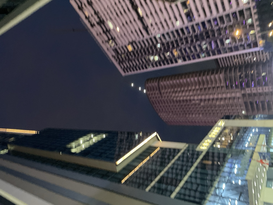 The image depicts a nighttime upward view of multiple high-rise buildings in Batumi, Georgia. On the left, a modern skyscraper with a dark glass facade exhibits reflective surfaces and horizontal strips of golden illumination. A sign displaying Georgian script, "ლუქს ორბი ჯგუფი" (Lux Orbi Group), is visible on its lower portion, above an entrance where a partially visible red and white flag, consistent with the Georgian national flag, is present. In the center, a tall, curvilinear building appears to be under construction or unfinished, characterized by exposed concrete structural elements and internal purple and yellow lighting on several levels. Above this central building, four small, bright white lights are aligned vertically in the dark blue night sky. To the right, a third high-rise building, primarily white, presents numerous illuminated windows and balconies displaying yellow, blue, and purple light. The overall scene is an urban environment at night. No individuals, specific actions, or interactions are discernible.