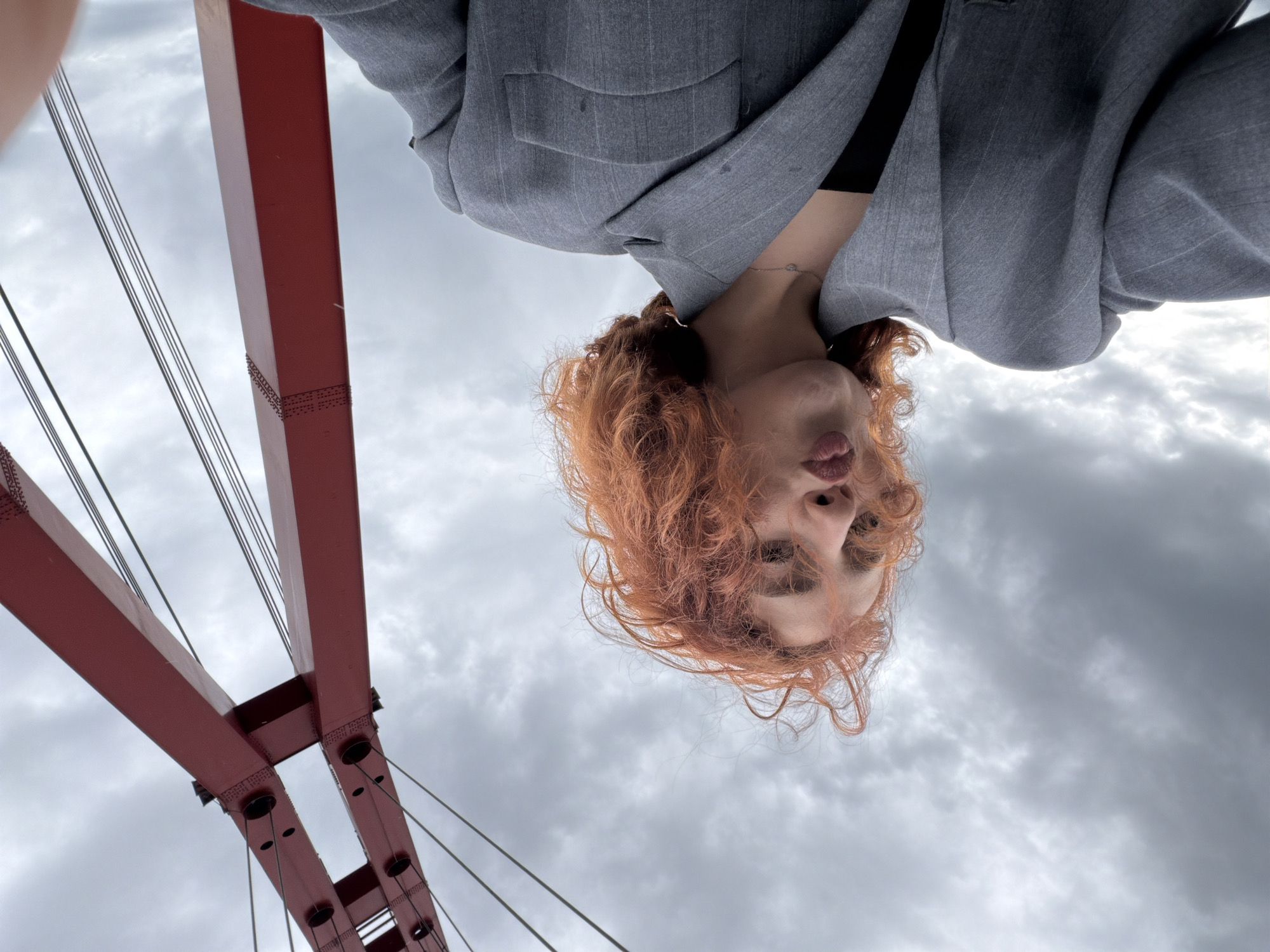 A low-angle portrait shows a person with red curly hair wearing a grey suit jacket. The person has light skin and is pouting. In the background, part of a red metal bridge structure is visible, and beyond that, a cloudy grey sky. Based on the bridge architecture, the location is likely Tbilisi, Georgia.