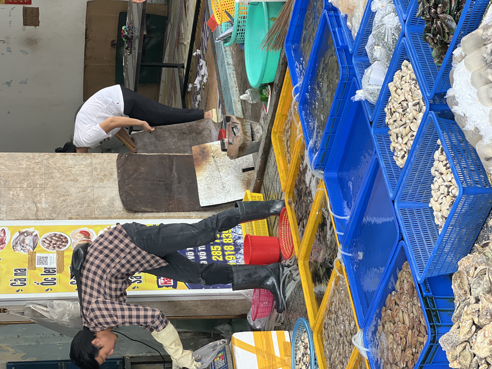 This image depicts a street-level seafood stall in Nha Trang, Vietnam. In the foreground, a man wearing a plaid shirt, black pants, and rubber boots is attending to a digital scale next to a yellow-taped white box. He is wearing protective gloves and appears to be preparing to weigh or package seafood. A red bucket and several red and yellow baskets are visible on the ground around him.
Behind the man, a vibrant yellow sign with Vietnamese text and images of seafood is prominently displayed. The sign includes a QR code and a phone number. Further back, a woman in a white t-shirt and black pants is seen from the side, interacting with a rustic clay stove.
The stall is filled with numerous blue and yellow plastic crates, many containing various types of seafood submerged in water. These include shellfish like clams and mussels, as well as what appear to be oysters. Some crates have water hoses running into them, likely for aeration. The ground is wet, indicating a typical market environment. The overall scene conveys a busy, functional marketplace setting in Nha Trang.
