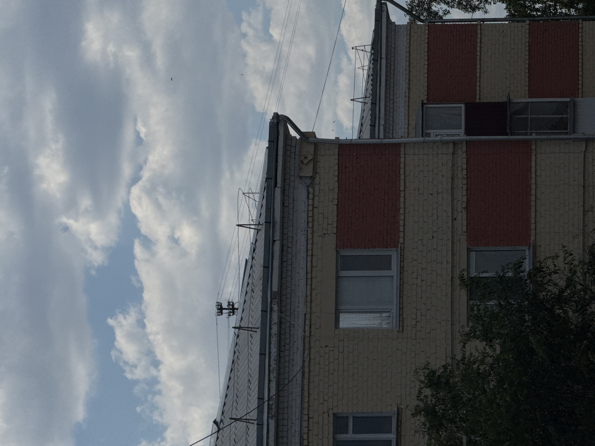 The image shows a close-up of the upper floors of two adjacent brick buildings, likely apartment blocks, in Zhovte, Ukraine. The building in the foreground has a facade of yellow brick with horizontal bands of red brick. It features several windows with light-colored frames. A grey metal roof with visible antenna structures and wires is visible at the top of the building. The building in the background, partially visible, shares a similar architectural style with yellow and red brickwork and a grey metal roof. The sky above is partly cloudy with white cumulus clouds against a light blue sky. A small dark object, possibly a bird or insect, is visible in the upper right portion of the sky. Trees with dark green foliage are visible in the lower left and right edges of the frame. The lighting suggests it is daytime, with the sun possibly behind the clouds or at an angle that creates soft shadows.