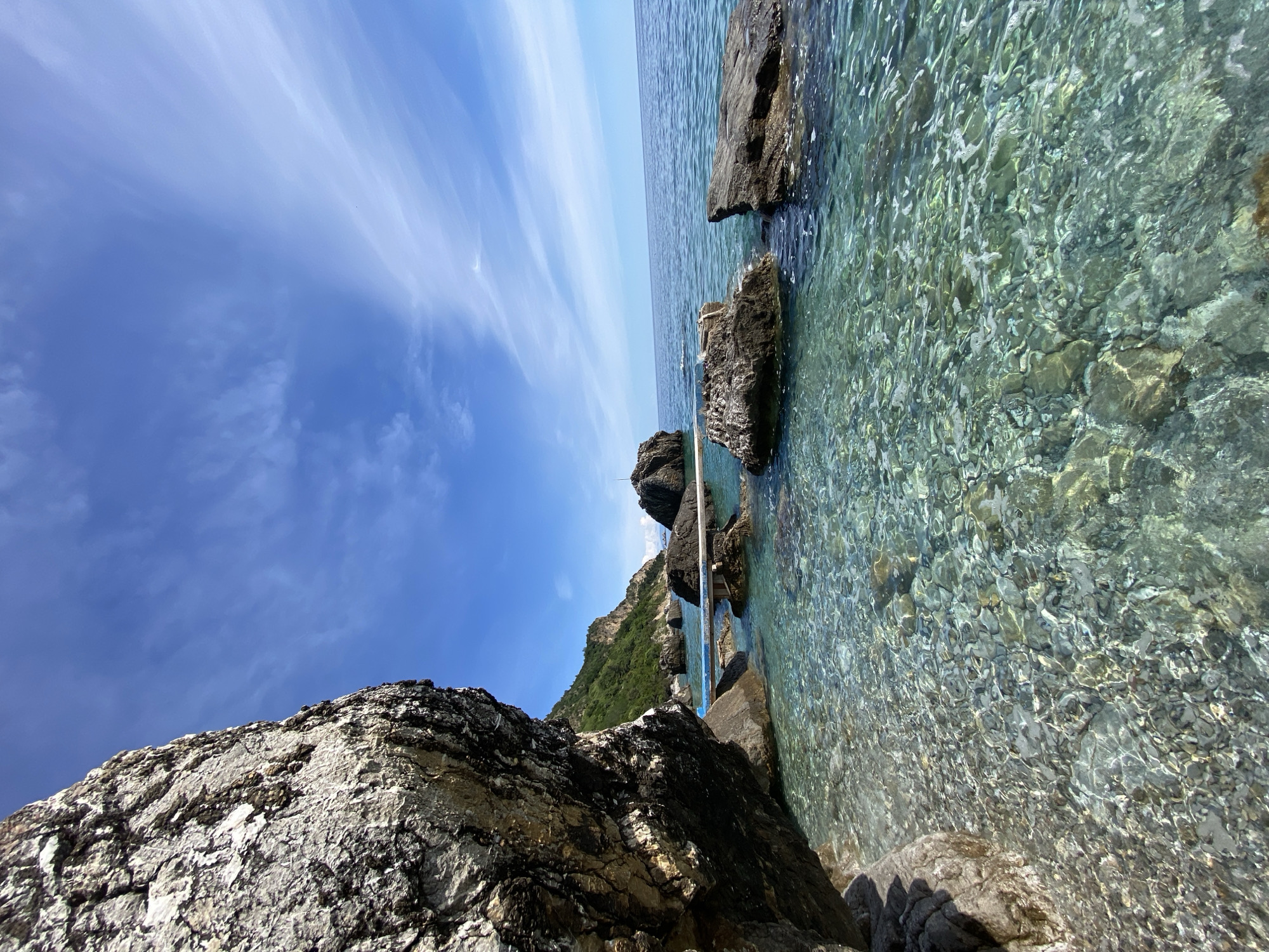 The image shows a rocky shoreline in Prijevor, Montenegro, with clear turquoise water lapping against the rocks. The sky is blue with wispy clouds. A rocky cliff is visible in the foreground, and some vegetation is visible on a hill in the middle ground. The image is taken from a low angle, giving a sense of immersion in the scene. A light-colored structure is partially visible in the water.