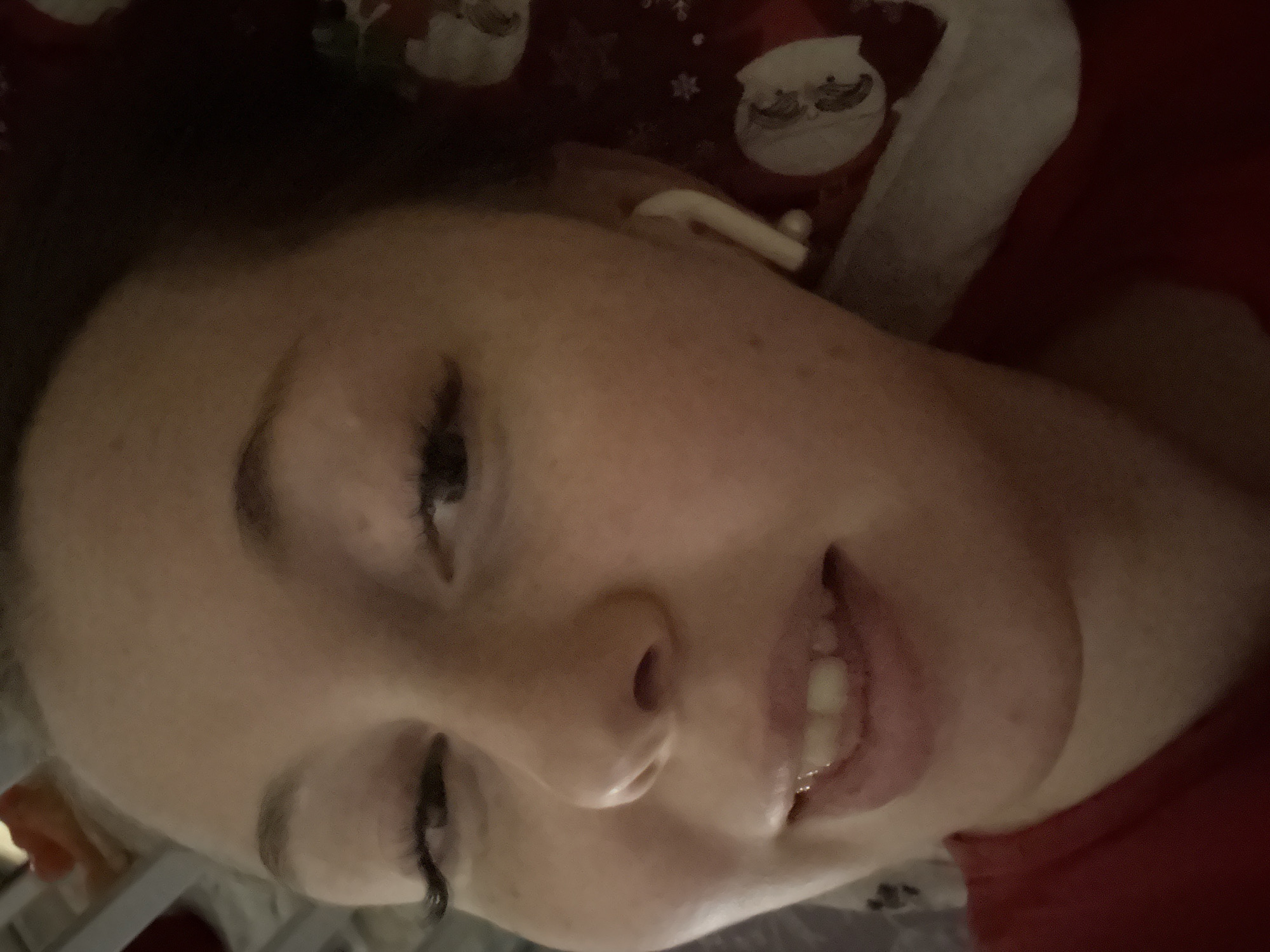 An upward-angled, close-up shot of a person, likely a woman, lying down indoors. The image is rotated approximately 90 degrees clockwise, showing the person's face tilted to the side. She has fair skin, dark eyelashes, and her mouth is slightly open, revealing her teeth. A white earbud is visible in her right ear. Her expression appears relaxed or sleepy. She is wearing a red garment, possibly pajamas or a top, patterned with festive white elements that resemble Santa Claus faces and snowflakes. The background is dark, suggesting low light conditions, possibly evening or night. No specific location cues other than an indoor setting are visible. There is no visible text in the image.