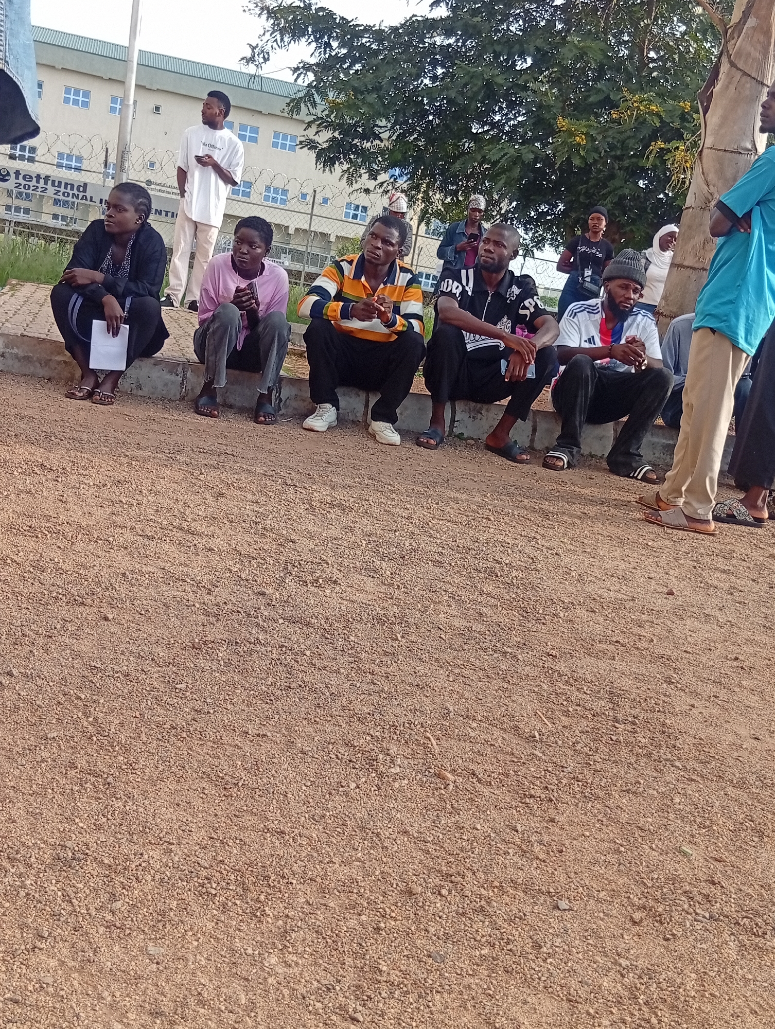 In Durumi, Nigeria, a group of people are seated on a low concrete ledge, facing forward. The individuals, appearing to be of various ages and genders, are dressed in casual attire. To the left, a woman in a black jacket and headwrap holds a document. Next to her, a woman in a pink shirt and dark pants looks towards the ground. To their right, a man in a striped shirt and dark pants sits with his hands clasped. He is followed by a man in a black and white shirt with lettering and dark pants, then a man in a white and blue soccer jersey and dark pants. The background reveals a modern building with multiple windows, surrounded by a wire fence. Trees with green foliage are visible above and behind the individuals. The ground in front of the ledge is covered in light brown gravel.