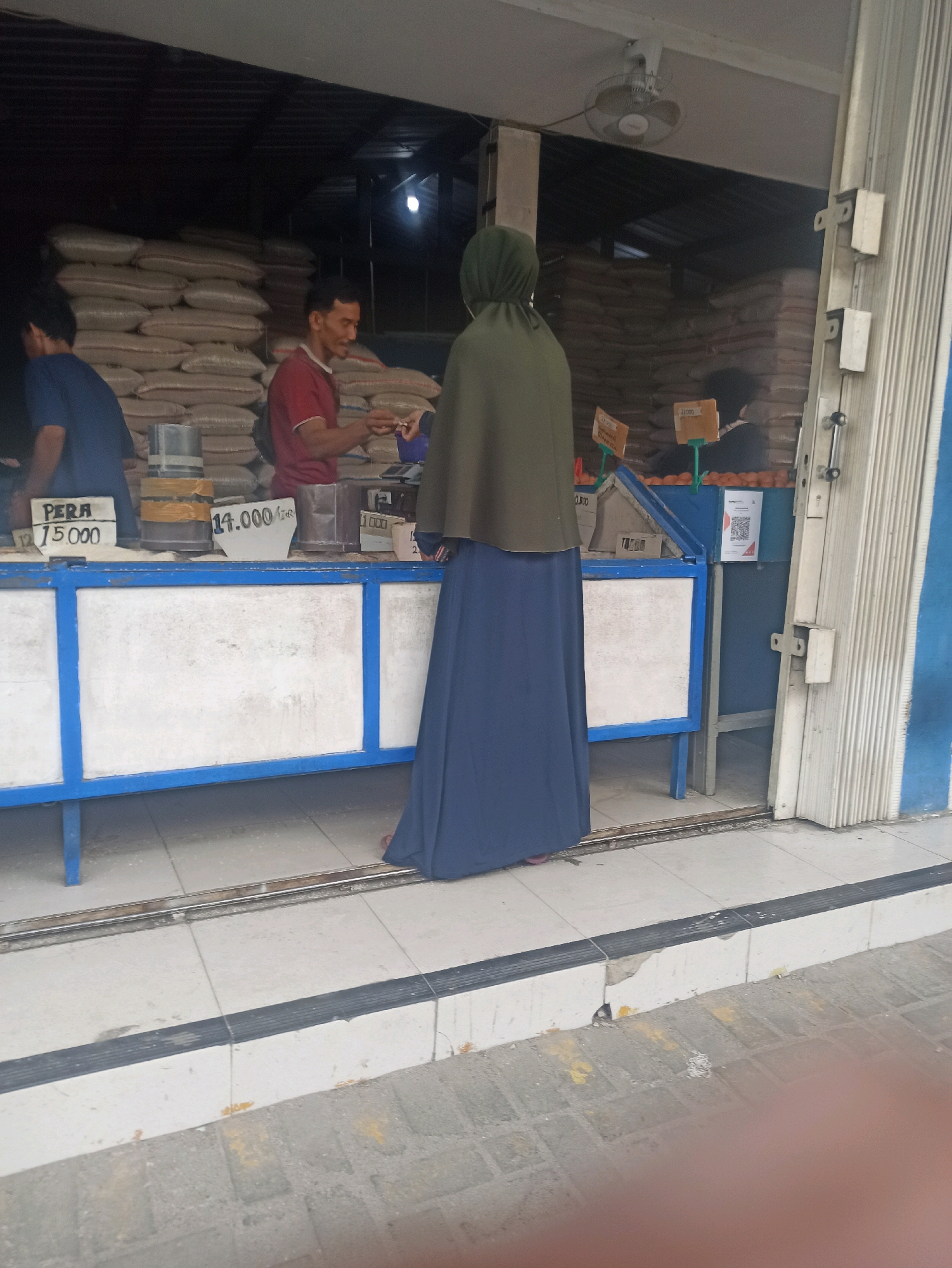 The image depicts an outdoor market scene in Telagamurni, Indonesia, likely at a food stall. In the foreground, a person wearing a navy blue long-sleeved shirt and dark pants stands with their back to the camera, partially obscured by the edge of a stall. A person in a maroon t-shirt is interacting with a customer, who is wearing a green hijab and a long navy blue skirt. The customer is holding a small item, possibly money or produce, and the vendor is reaching out to take it or hand something over.
The stall itself is a long, blue and white counter. Various items are displayed on the counter, including price tags with Indonesian currency. One tag reads "PERA 15.000", and another says "14.000 GR". There are also containers holding goods, one with a yellow band. Behind the stall, large sacks of what appear to be rice or grain are stacked high, filling the background. A ceiling fan is visible above the stall, and a partially rolled-up metal shutter door is on the right side of the frame. The ground is paved with light-colored square tiles, with a darker strip running along the edge of the stall. The overall atmosphere suggests a busy market day.
