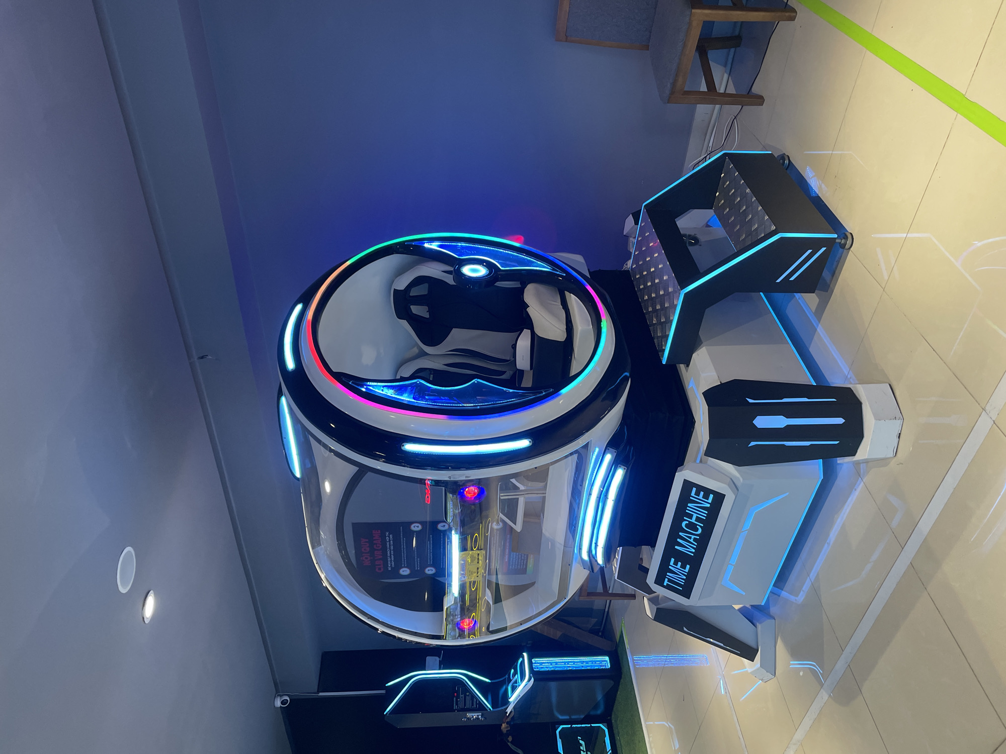 The image shows a "Time Machine" VR game arcade machine in Nha Trang Ward, Vietnam. The machine features a spherical, transparent dome with colorful neon lights. Inside the dome are two seats. The machine has a control panel on the right with the words "TIME MACHINE" lit up. The floor is tiled. In the background, there is a chair and part of a wall.