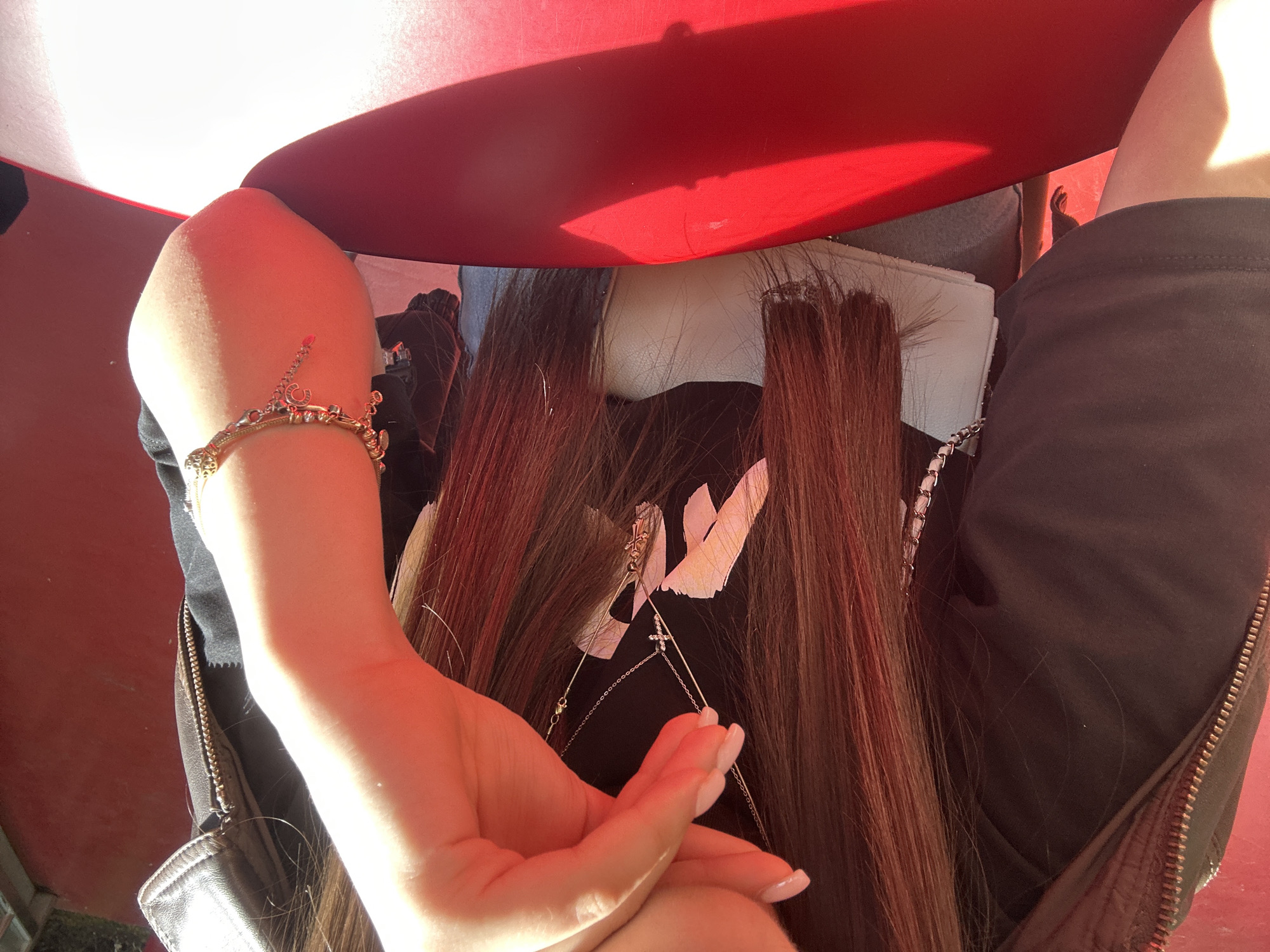 A close-up, angled view shows a person wearing a black shirt with a white design, seated beneath a red umbrella. The person's left arm is visible, adorned with a gold bracelet. Long, brown hair is draped down the person's back, partially obscuring the shirt's design. The person's right hand is brought up to touch the left hand. The setting is unknown and cannot be verified to be the city of Yerevan, Armenia.