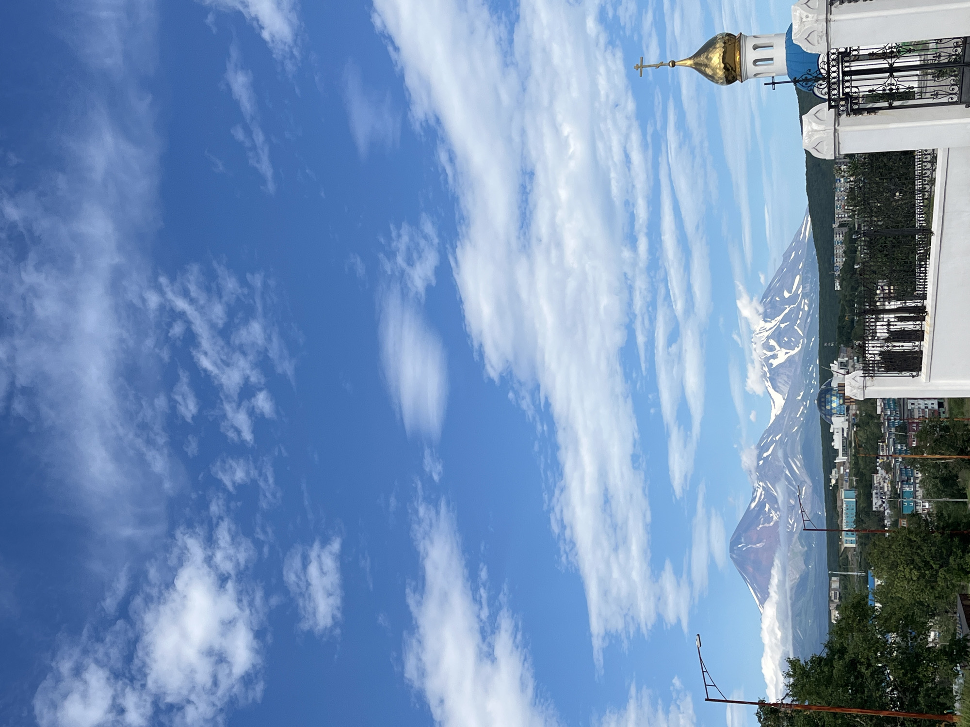 The image depicts a panoramic view of Petropavlovsk-Kamchatsky, Russia, with several prominent volcanoes in the background. In the foreground on the right, a white fence with decorative elements and black metal gates encloses a structure topped with a golden dome and a cross, characteristic of an Orthodox church. A smaller black cross is visible on a blue section of the church's roof. To the left, a street lamp with a single light is positioned near a cluster of green trees. In the mid-ground, a cityscape with various buildings, including some with blue and teal facades, stretches across the horizon. Behind the city, snow-capped volcanoes rise majestically, with the largest appearing reddish-brown at its peak. Wisps of clouds cling to the volcanic slopes. The sky above is a vibrant blue, dotted with scattered white cumulus clouds. The overall scene suggests a clear day with a dramatic natural landscape juxtaposed with urban development and religious architecture.