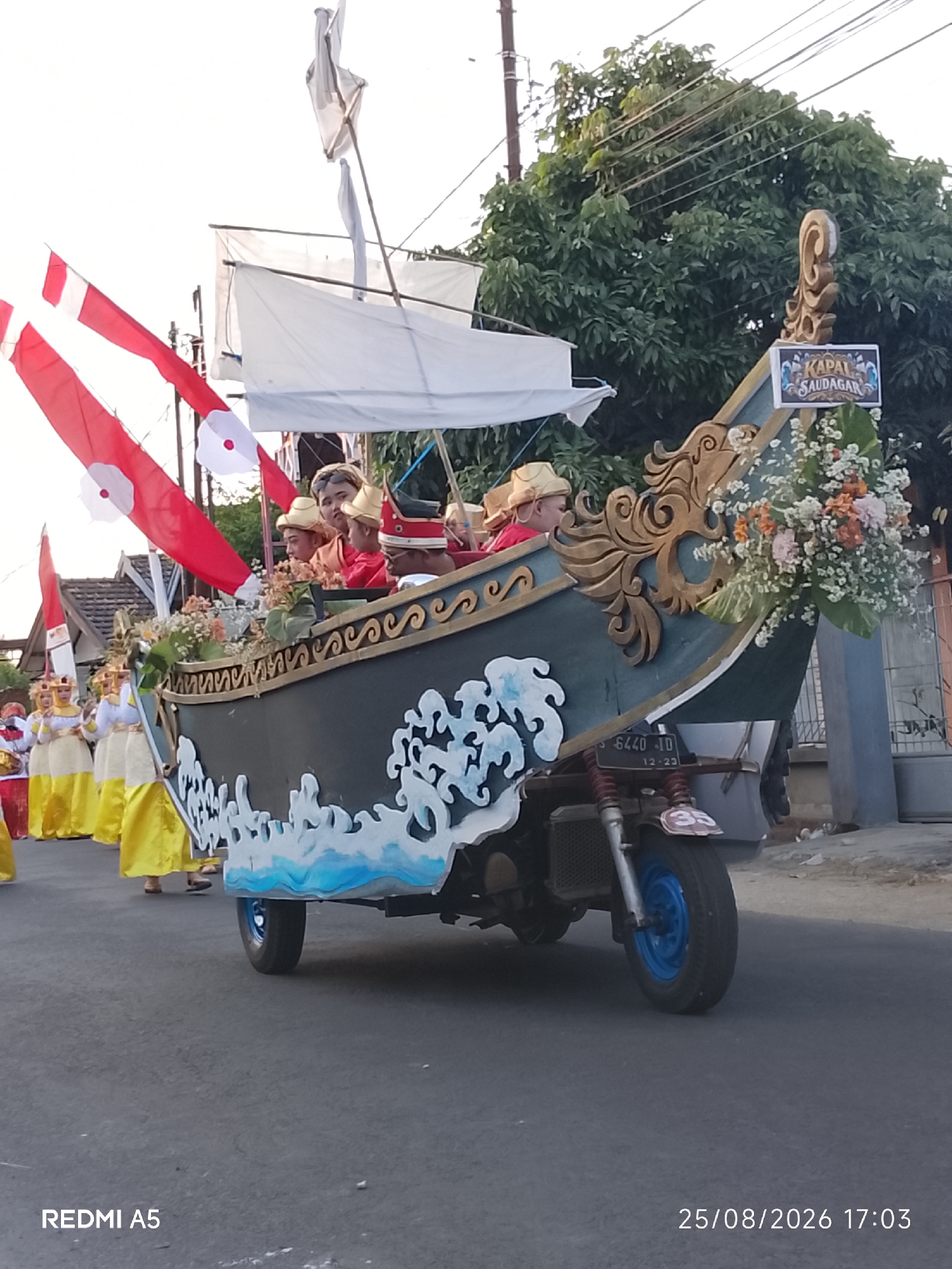 In Semanding, Indonesia, a colorful parade float resembling a ship is being transported, likely by a modified motorcycle chassis visible at its base. The float is adorned with red and white flags, white sails, and decorative elements in gold and blue. Several individuals, some in traditional attire and hats, are seated within the float. In the background, lush green trees and buildings suggest a residential or commercial area. The scene is captured during daylight hours, with a visible timestamp indicating August 25, 2026, at 17:03.