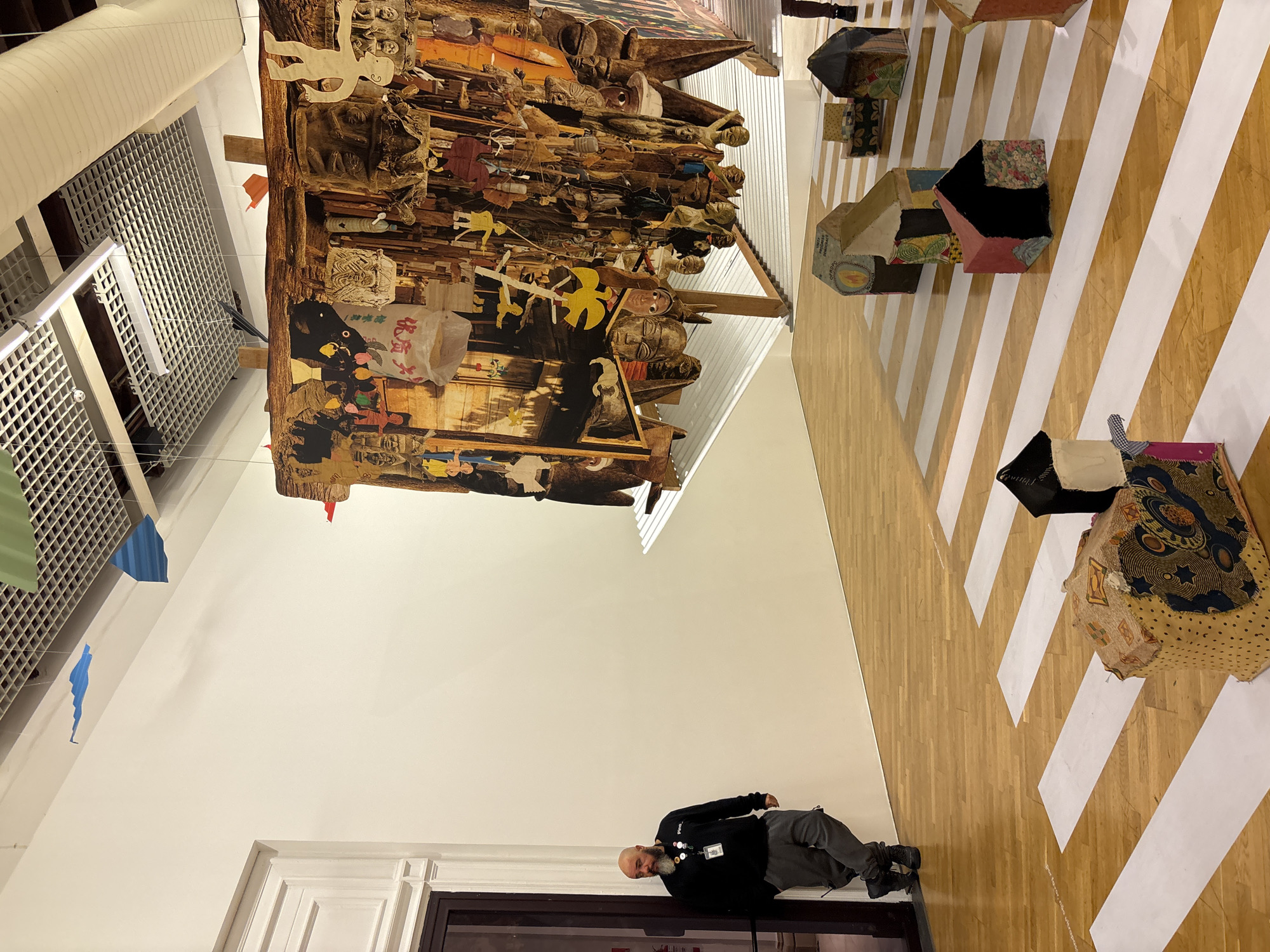A brightly lit interior of an art gallery is viewed from a slightly oblique angle. Dominating the scene is a large, complex hanging tapestry or assemblage of various wooden figures and objects. Below, a man dressed in dark clothing lies on the floor next to a doorway. The floor is covered with a pattern of parallel white stripes. A series of three-dimensional patchwork objects are arranged on the floor, seemingly part of the exhibit. The upper part of the gallery features architectural details like a white lattice ceiling and white walls. Given the context of the image, it is likely the location is an art gallery in the city of São Paulo, Brazil. The interaction is the man observing the artwork.