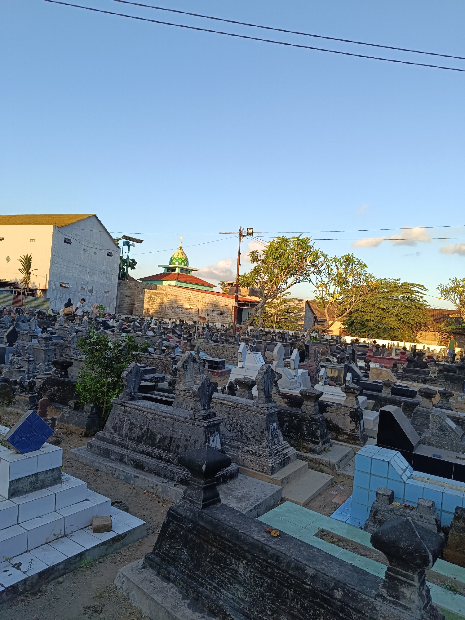 A medium shot captures a sun-drenched cemetery in Banguntapan, Indonesia, under a clear blue sky with a few scattered clouds. The foreground is dominated by a dense arrangement of tombstones, varying in size, shape, and material, from weathered grey stone to lighter colored, possibly marble, structures. Some tombstones are simple rectangular slabs, while others are more elaborate, featuring decorative carvings and tiered bases. A few graves are adorned with blue and green tiles. In the mid-ground, a mosque with a green dome and red roof stands prominently, its minaret visible. Behind the mosque, a multi-story building with a yellow-tiled roof and grey walls is partially visible. Several trees, some with leafy canopies and others sparsely branched, are scattered throughout the scene, casting shadows on the tombstones. A few people, dressed in casual attire, are present, scattered throughout the cemetery. One group of three individuals is seen conversing near the building in the mid-ground. Another individual, wearing a green shirt, stands in the far right mid-ground. The ground is a mix of dirt and patches of sparse vegetation. Power lines are visible traversing the upper portion of the image. The overall impression is one of a peaceful, albeit somber, resting place bathed in the warm light of what appears to be late afternoon.