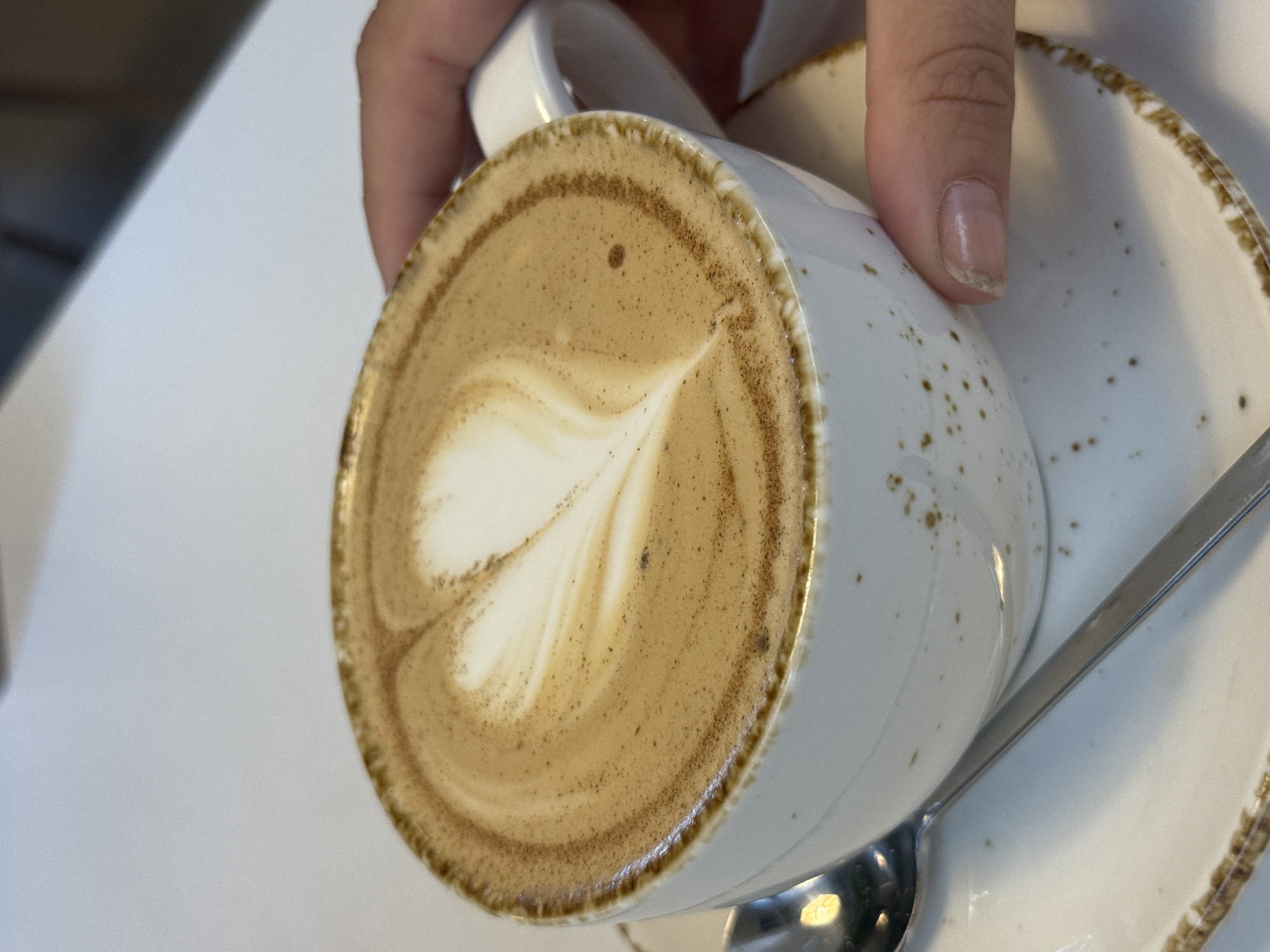 A close-up shot shows a white ceramic mug with a latte art heart design, filled with coffee and topped with foam. The mug is speckled with brown dots and has a brown rim. It rests on a matching saucer. A silver spoon is partially visible, lying on the saucer to the left of the mug. A hand, with a thumb and index finger, is holding the mug, with the thumb resting on the saucer and the index finger on the side of the mug. The background is a plain, light-colored surface, likely a table. The scene takes place indoors. There is no information in the image to suggest the location is in Naberezhnye Chelny, Russia.