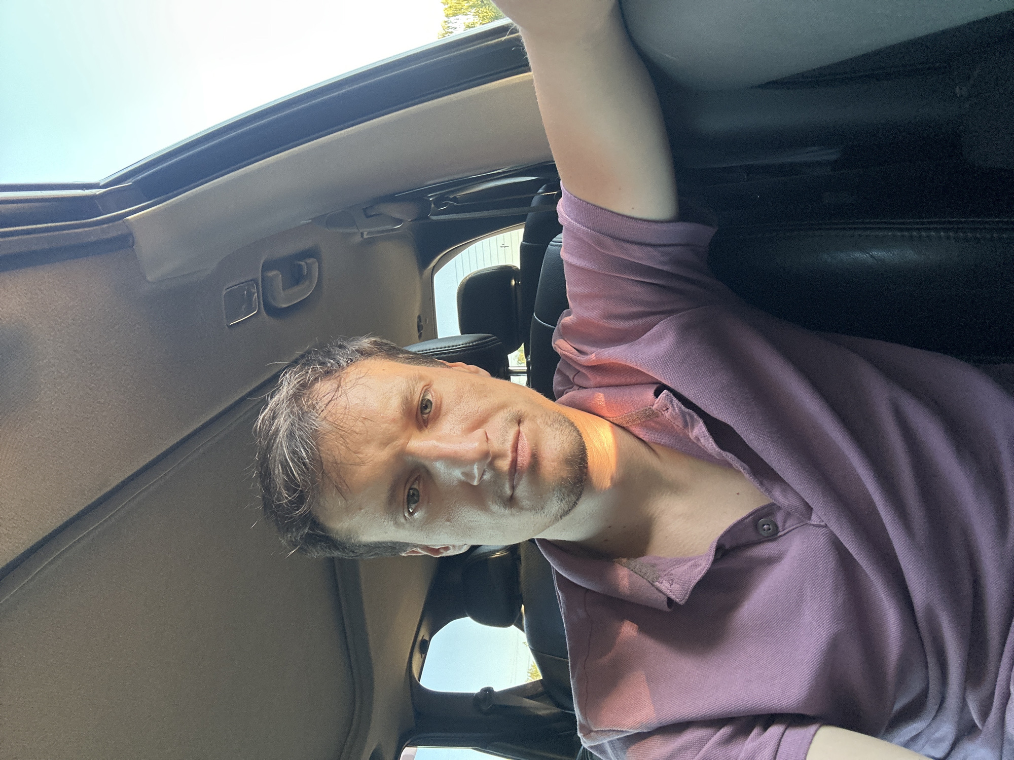 A medium shot captures a man in a purple polo shirt sitting in the driver's seat of a car. The man has short, light brown hair with some graying at the temples and a light beard. He is looking directly at the camera with a neutral expression. The interior of the car is visible, including the car's sunroof, headliner, and dashboard. The background shows the sky and some trees, suggesting an outdoor setting. The lighting indicates it is daytime, possibly late afternoon due to the warm, angled light on the man's face. The car appears to be parked, and the scene is composed as a selfie. There is no information available to determine the specific location in the city of Makiv, Ukraine, from the image itself.