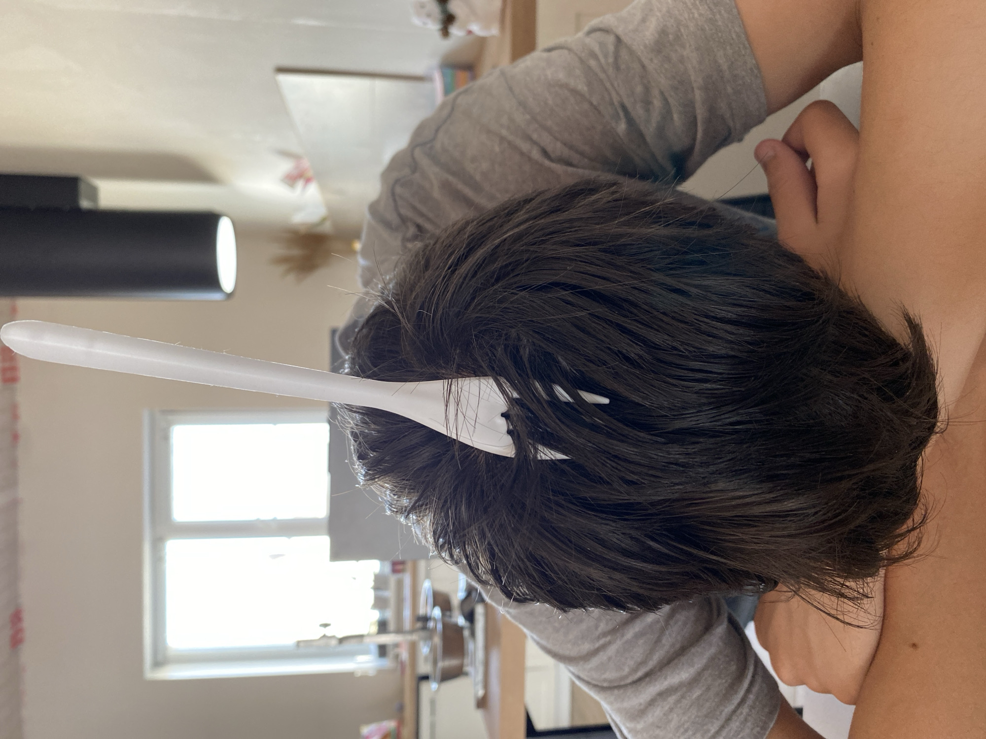 A person is seen from an overhead perspective, lying down. A white plastic fork is stuck into the person's dark hair. The person is wearing a gray long-sleeved shirt. A light source, possibly a lamp, is visible on the left. A window is visible with bright light coming through it. The scene appears to be indoors. There is no information to identify the location as Chaikovsky, Russia.