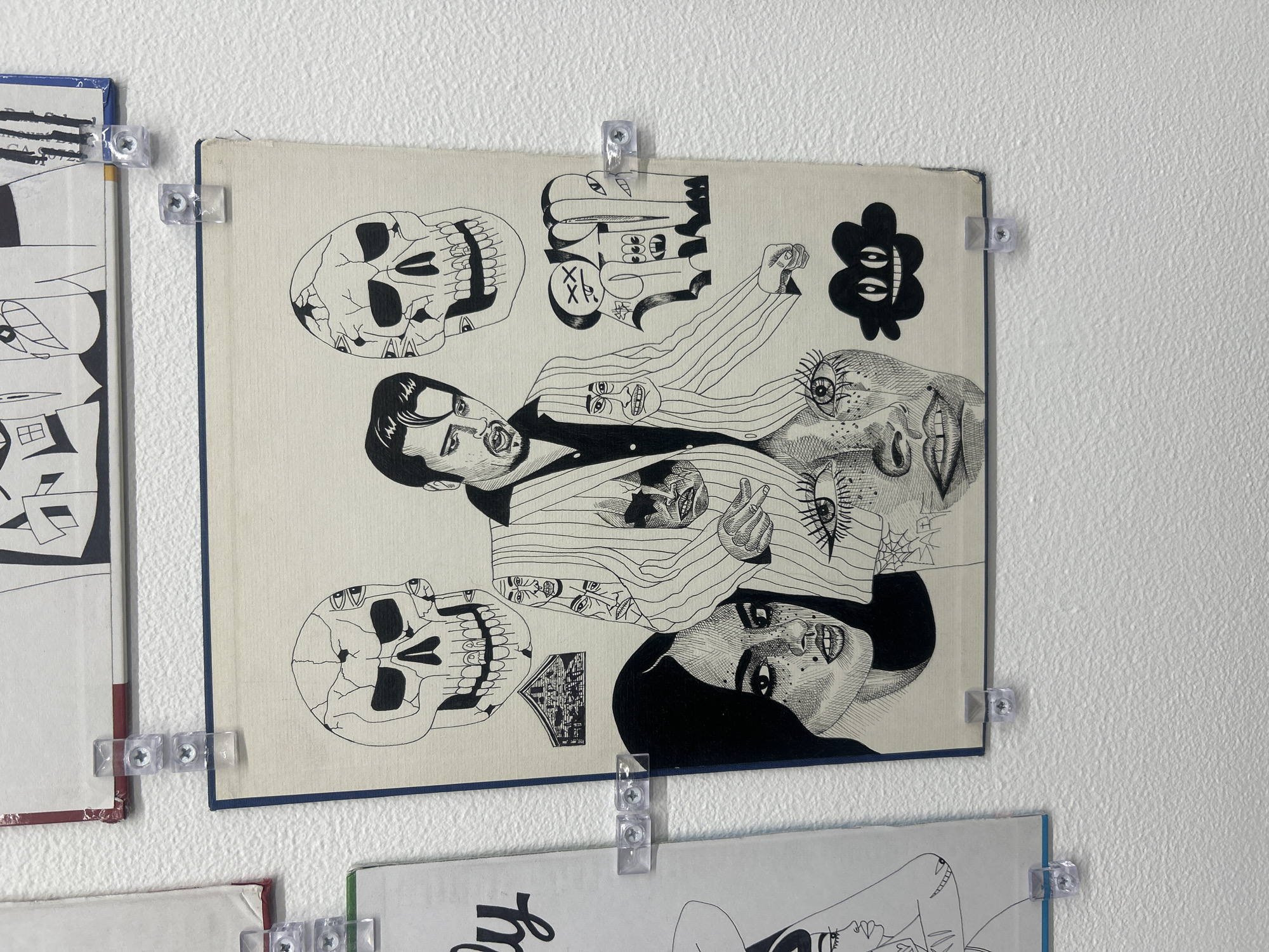 The image shows a collection of framed black and white drawings mounted on a white wall. The drawings feature diverse subjects, including skulls, faces with exaggerated expressions, and abstract designs. The frames are simple, with a narrow blue border. The drawings are attached to the wall using clear plastic clips with screws. The background is a plain white wall with a slightly textured surface. The location is indoors, but specific location in Los Angeles, United States of America is not discernible from the image. There are no people visible in the image, only the drawn representations.