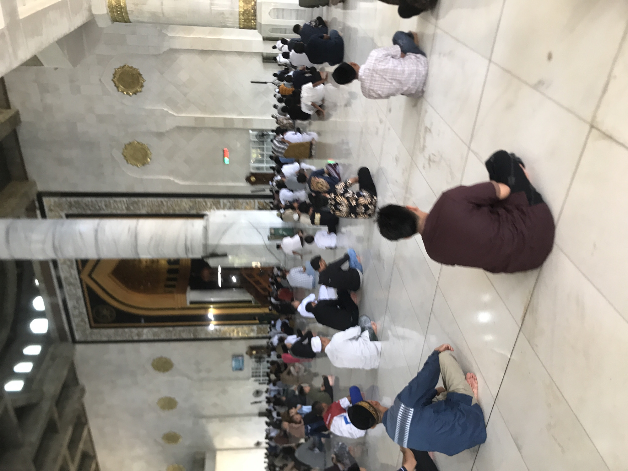 The image shows a group of people praying inside what appears to be a mosque in Lhokseumawe, Indonesia. The people are sitting on the floor in rows. The interior of the mosque has white tiled floors and columns, with decorative gold elements on the walls and ceiling.