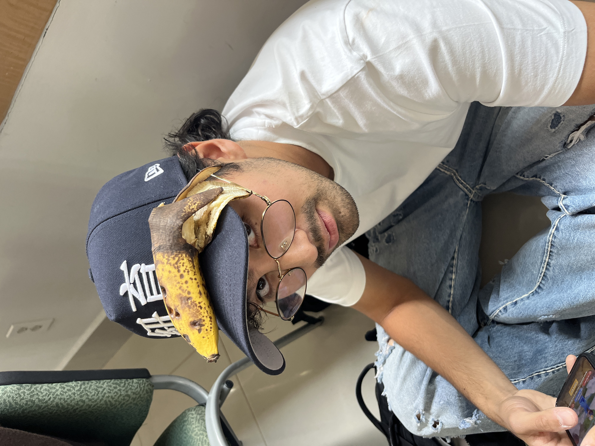 The image, rotated 90 degrees clockwise, captures a young man seated indoors in San Andrés Cholula, Mexico. He is looking directly at the camera with a neutral, slightly inquisitive expression. He wears a white short-sleeved t-shirt, light blue ripped jeans, and a dark blue baseball cap. Perched humorously on his cap is a ripe, partially peeled banana with brown spots.
The man has dark hair, pulled back, and wears round, gold-rimmed glasses. He has light stubble on his face. His left arm is visible, holding a smartphone that displays a colorful screen, likely a mobile game or application featuring a blue figure.
The setting appears to be an indoor environment, possibly a public space or waiting area. A chair with green, textured upholstery and a metal frame is visible to the left. The background consists of a plain, light-colored wall. White Asian characters are embroidered on the front of his baseball cap. The overall mood of the image is informal and lighthearted.