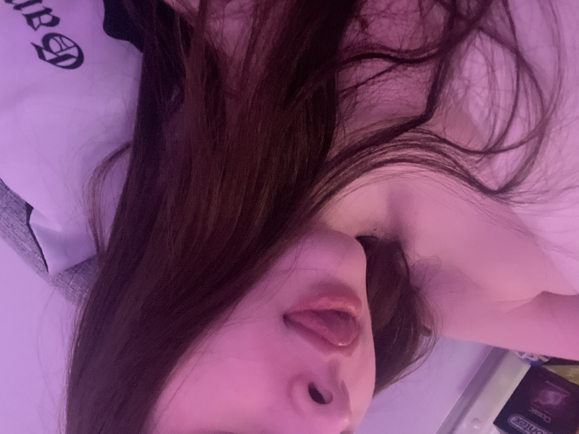 A close-up, rotated image displays a person with long, dark hair. The person's face is angled, revealing their nose, mouth with the tongue sticking out, and part of their neck. The image is bathed in a pink or purple hue. The background shows a pillow with dark graphics and what appears to be a box of condoms. The location is not identifiable within the image.