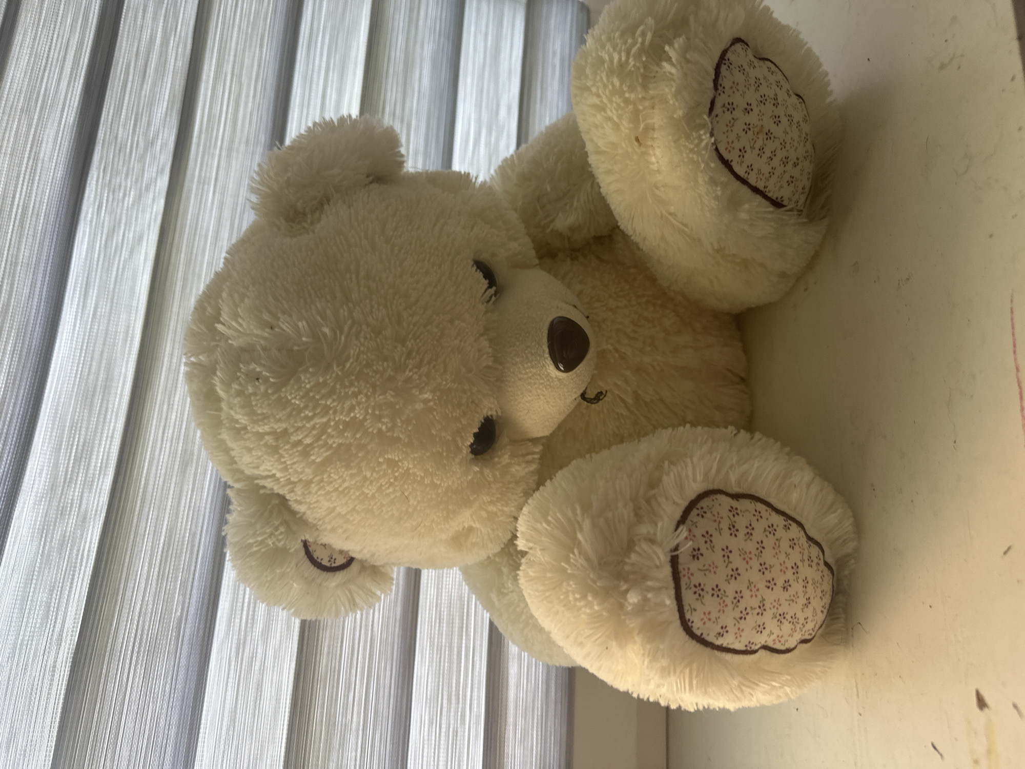 A fluffy, cream-colored teddy bear is seated on a white surface in front of a horizontally striped window blind. The teddy bear is facing forward, with its head tilted down slightly and its eyes looking downward. Its paws are resting in its lap, and its feet are placed in front of it. The soles of its feet have a light-colored fabric insert with a small floral pattern. The background consists of the window blind, which is composed of alternating opaque and sheer horizontal bands, creating a striped effect. The lighting appears to be artificial and casts a warm glow on the teddy bear and the surface it is on. The image does not provide any information about its location in Maladzyechna, Belarus, or any people present.