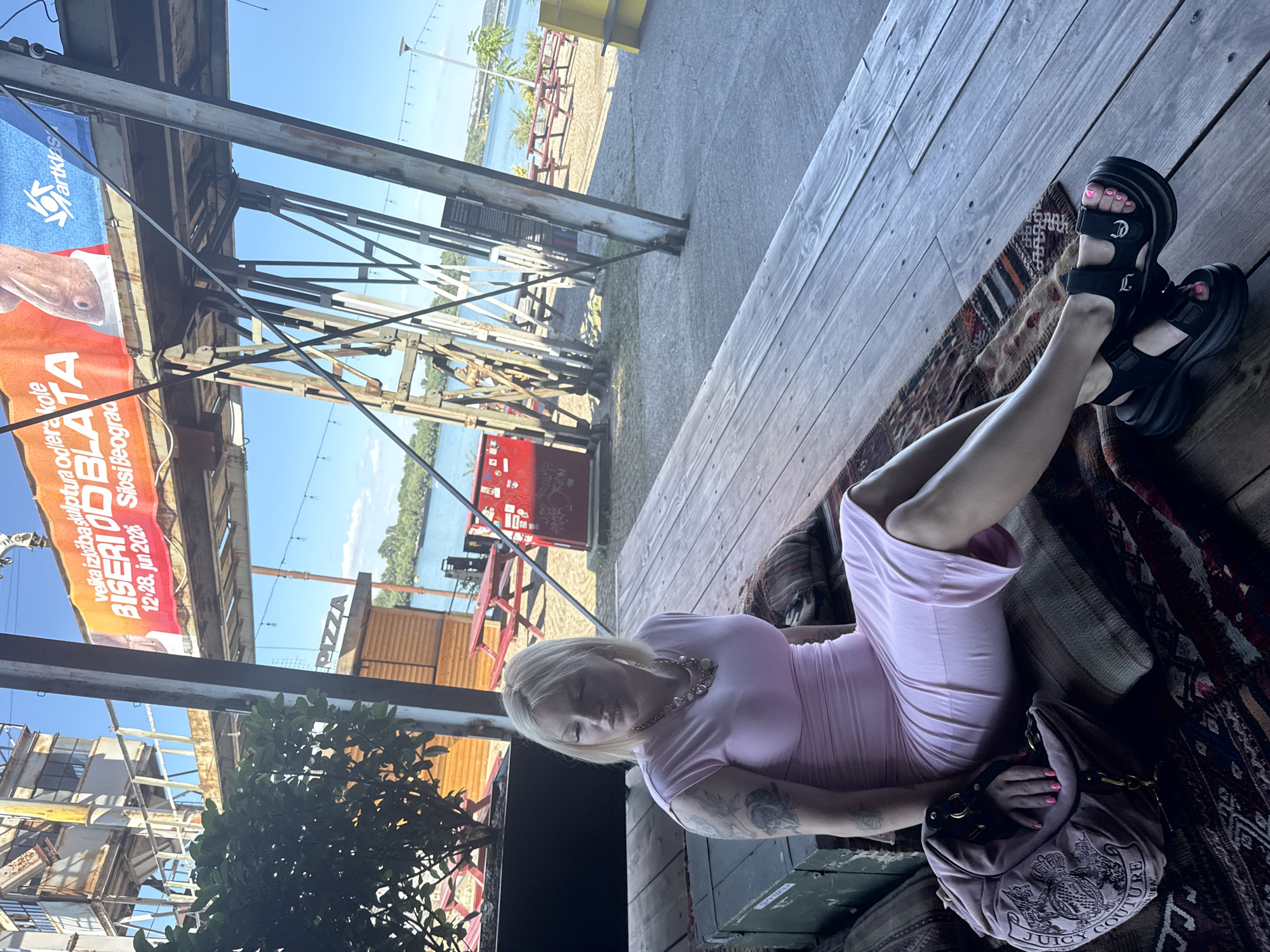 A blonde woman is sitting on a wooden platform, outdoors in what appears to be a recreational area. She is wearing a pink dress and black sandals. A pink purse with the "Juicy Couture" logo is beside her. Behind her, there is a river with trees and buildings on the opposite bank. A large banner with Serbian text and an image of a fish is visible in the background, likely advertising an event. The scene suggests a relaxed outdoor setting, possibly a waterfront venue in Belgrade, Serbia, given the visible signage.