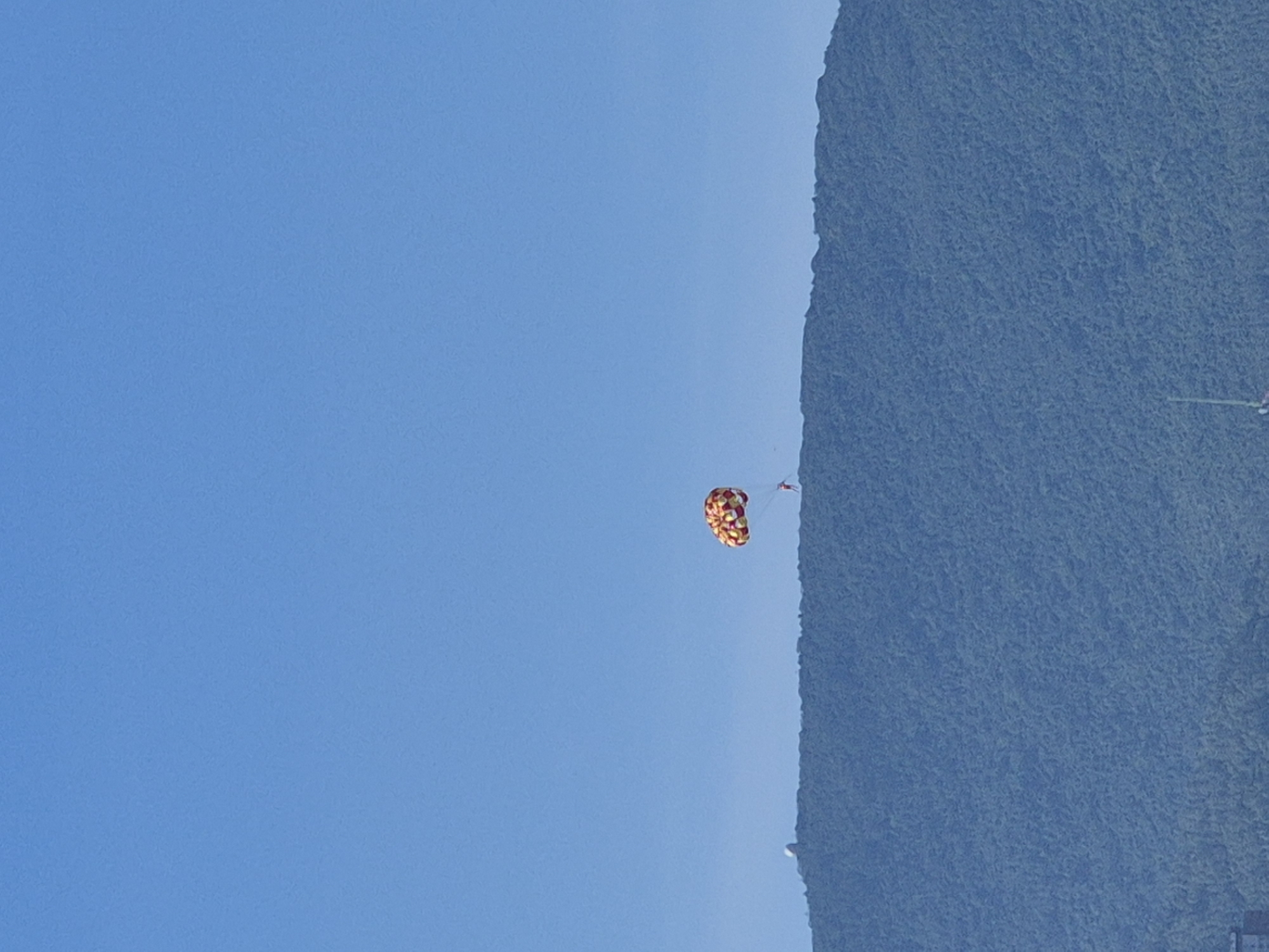 The image shows a parasailer in the sky above a mountain range in Đà Nẵng, Vietnam. The parasail is colorful, with red, yellow, and white patterns. The parasailer, dressed in red, is suspended beneath it. A white dome-shaped structure, possibly an observatory or weather station, is visible on the mountain summit to the left. The mountain is covered in lush green vegetation. The sky is a clear, bright blue. In the distance, at the bottom of the image, faint outlines of buildings and what appears to be a boat or mast suggest a coastal or harbor area.