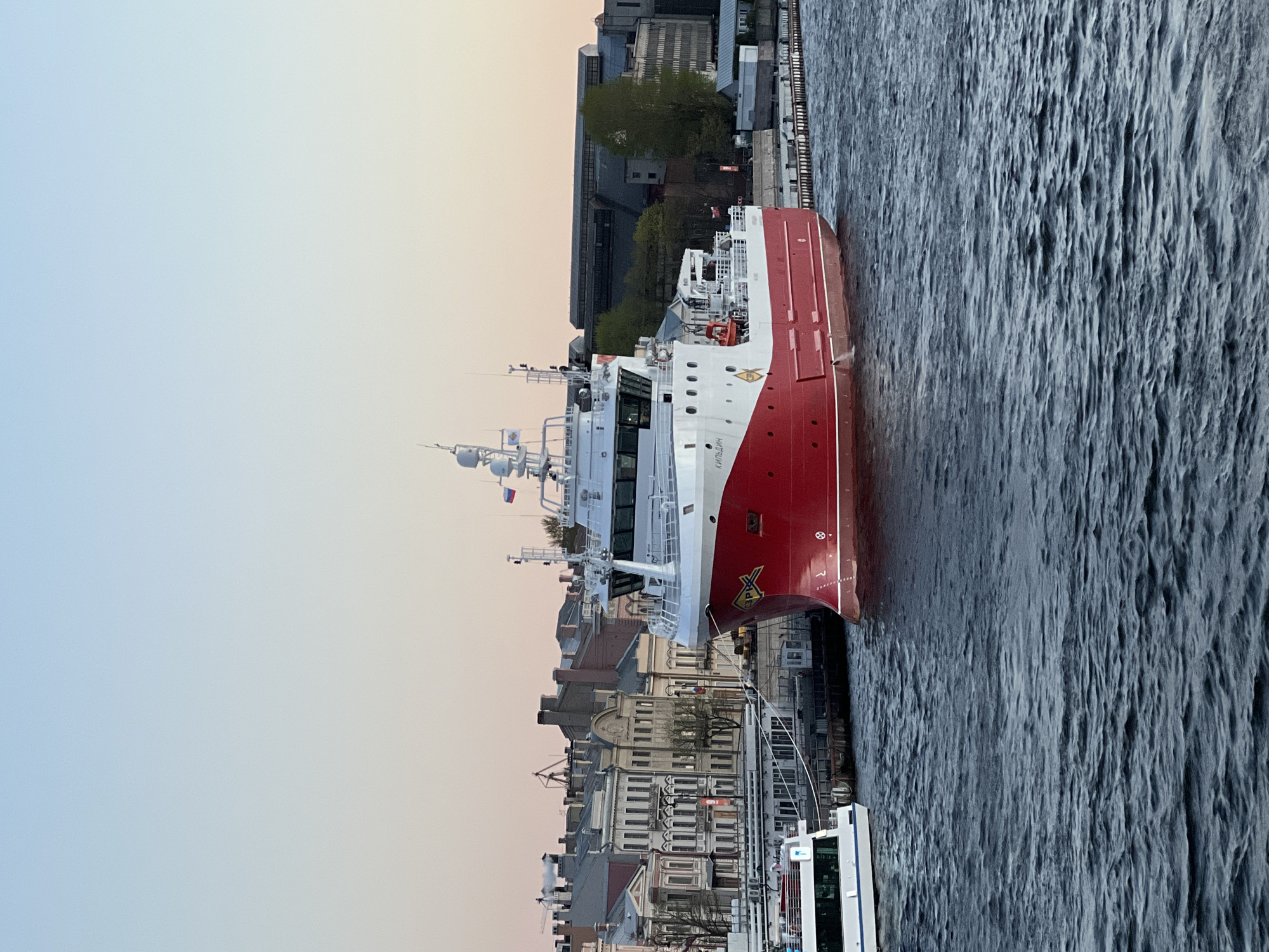A red and white ship, identified as the research vessel "Akademik Mstislav Keldysh" based on lettering on its side, is docked alongside a pier in Saint Petersburg, Russia. Several buildings line the waterfront, including what appears to be a mix of older, multi-story structures with detailed facades, and a more modern, industrial-looking building further back. The sky is a pale gradient of blue and orange, suggesting either sunrise or sunset. The water shows surface texture, indicating wind or currents. A second, smaller white vessel is visible near the foreground.