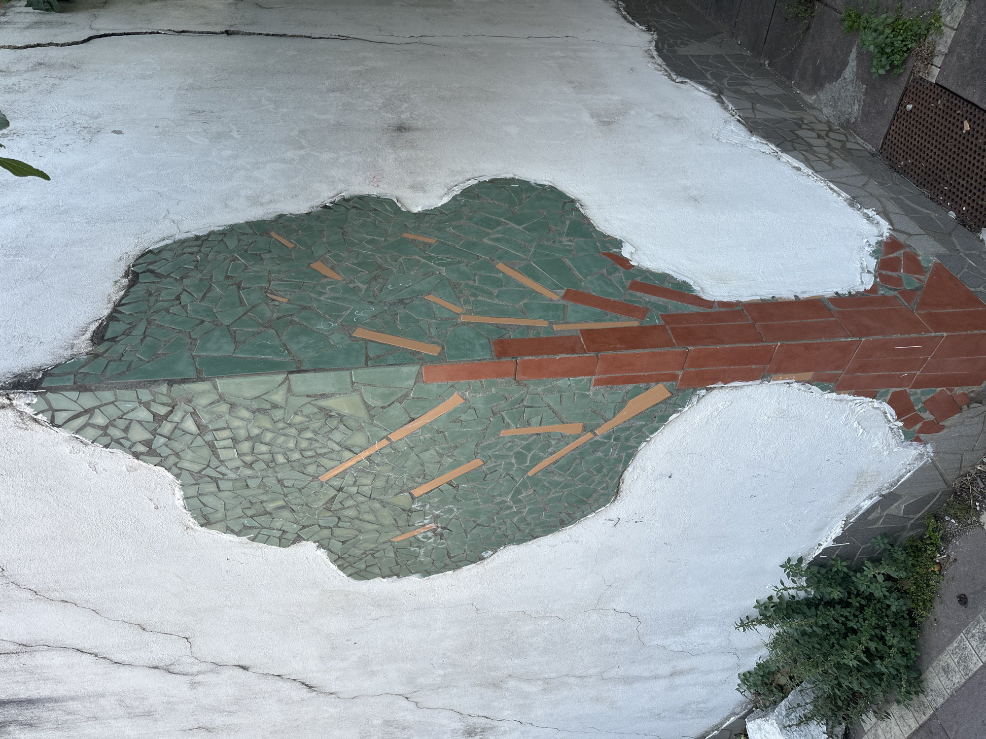 An overhead shot shows a concrete surface partially covered with a mosaic in Budva, Montenegro. The mosaic depicts a stylized form, possibly an abstract tree, with green tile fragments forming the canopy and red brick sections suggesting a trunk and branches. Irregular pieces of light tan tile are interspersed within the green mosaic. The mosaic is surrounded by white concrete, which appears to be cracked and aged. Patches of greenery are visible at the edge of the concrete surface. A grated metal cover is situated in the upper right corner. The overall impression is of a decaying public space with an integrated art piece.