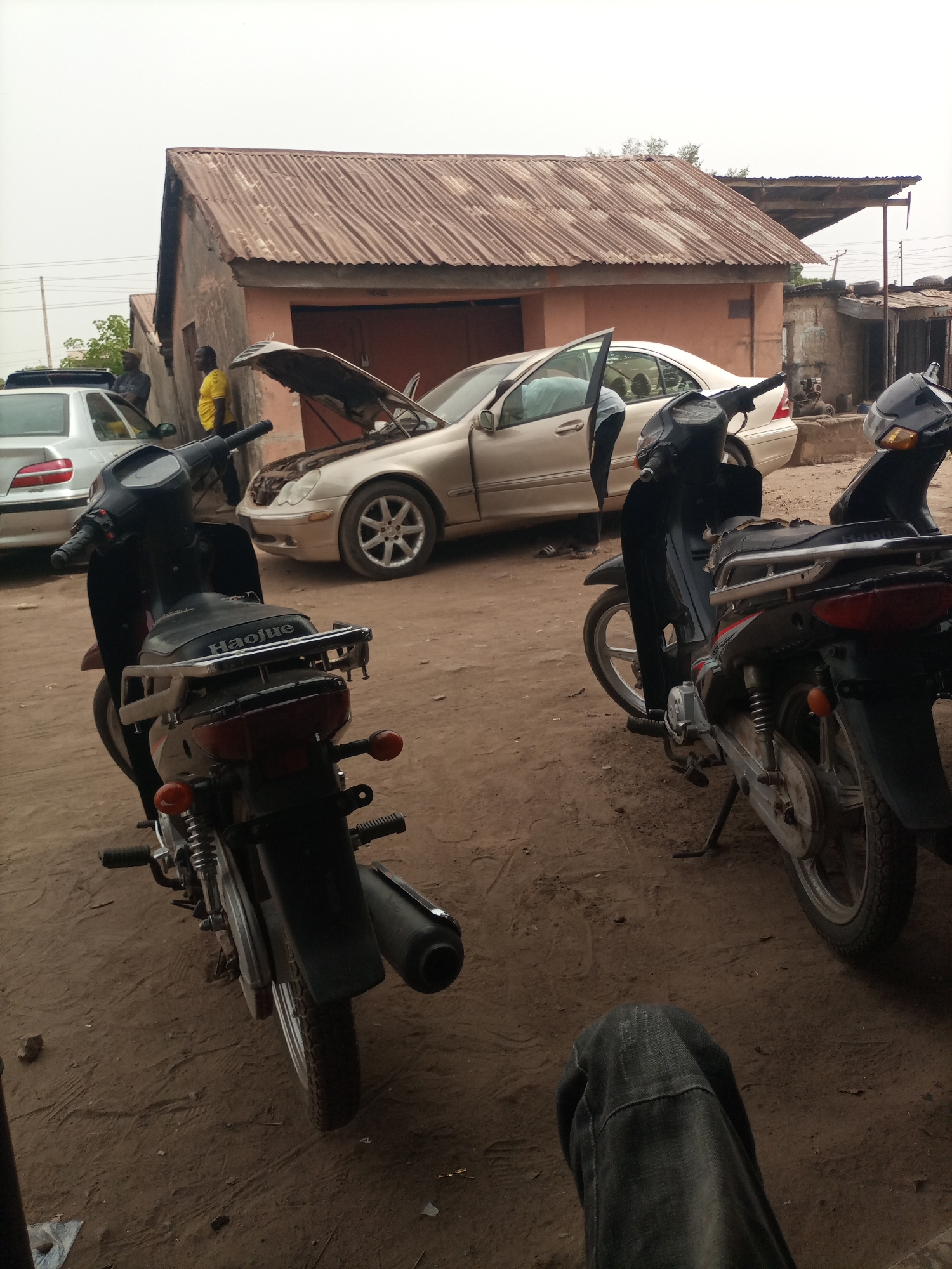 The image shows a dusty outdoor auto repair scene in Barden Yana, Nigeria. Two black motorcycles are parked in the foreground. Behind them, a gold Mercedes-Benz has its hood open; the driver's side door is also ajar. To the left, a silver sedan is partially visible. Two men stand near the vehicles; one wears a yellow shirt. A low, rust-colored building with a corrugated metal roof is behind the cars. The ground is dirt. A person's legs, clad in dark jeans, are visible in the lower portion of the frame.