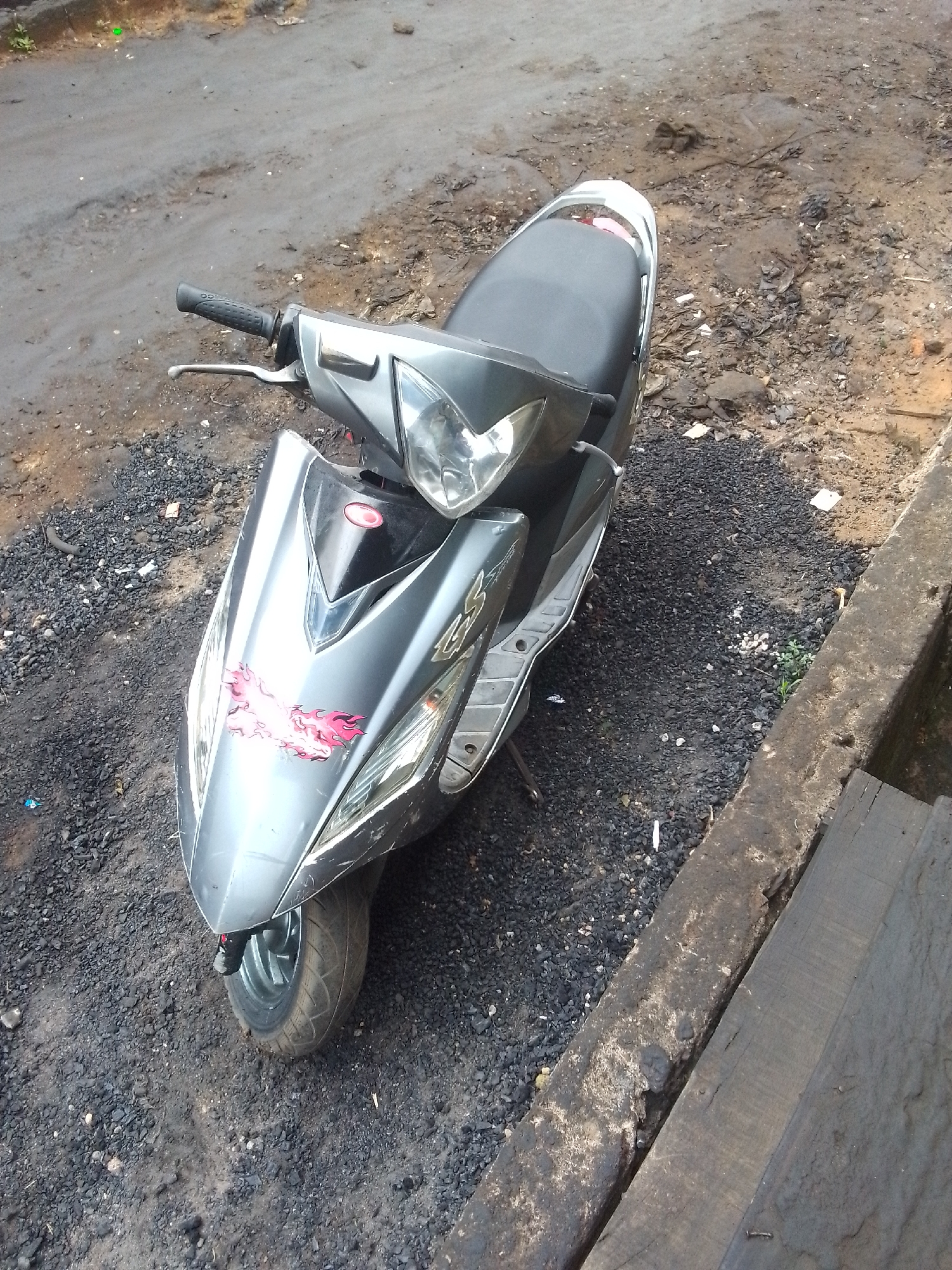 A gray motor scooter is parked on a surface of mixed gravel, dirt, and asphalt. The scooter has red flame decals on its front fender. The surrounding area appears to be a street edge or roadside with a concrete curb and a narrow strip of pavement. The location is in the city of Onitsha, Nigeria.