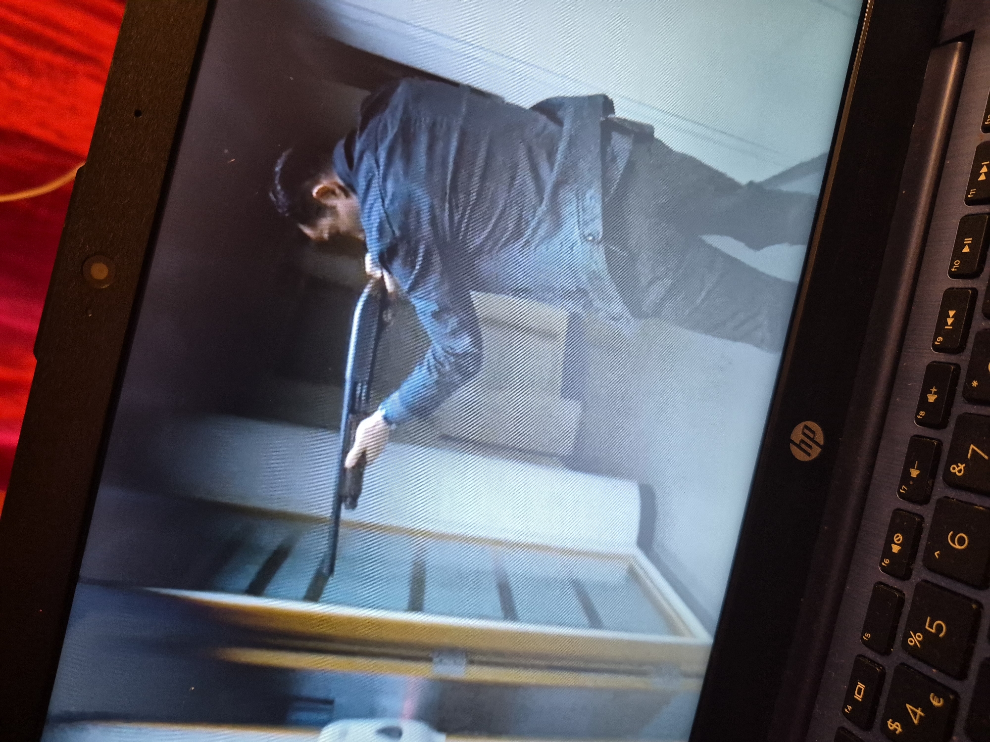 A man is holding a shotgun in a hallway. The man is wearing a dark jacket and dark pants. The hallway appears to be inside a building, possibly an office or institutional setting, with doorways and walls visible. The image is captured on a laptop screen, as indicated by the laptop keyboard in the foreground. There is no indication of the location being in Sandwell, United Kingdom.