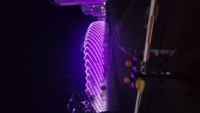 A building is illuminated with bright purple lights at night. The lights form a series of horizontal lines that curve along the building's exterior. A road and a gate arm with yellow lights are visible to the right of the building. Other buildings can be seen in the background. The lighting is very bright against the dark night sky.