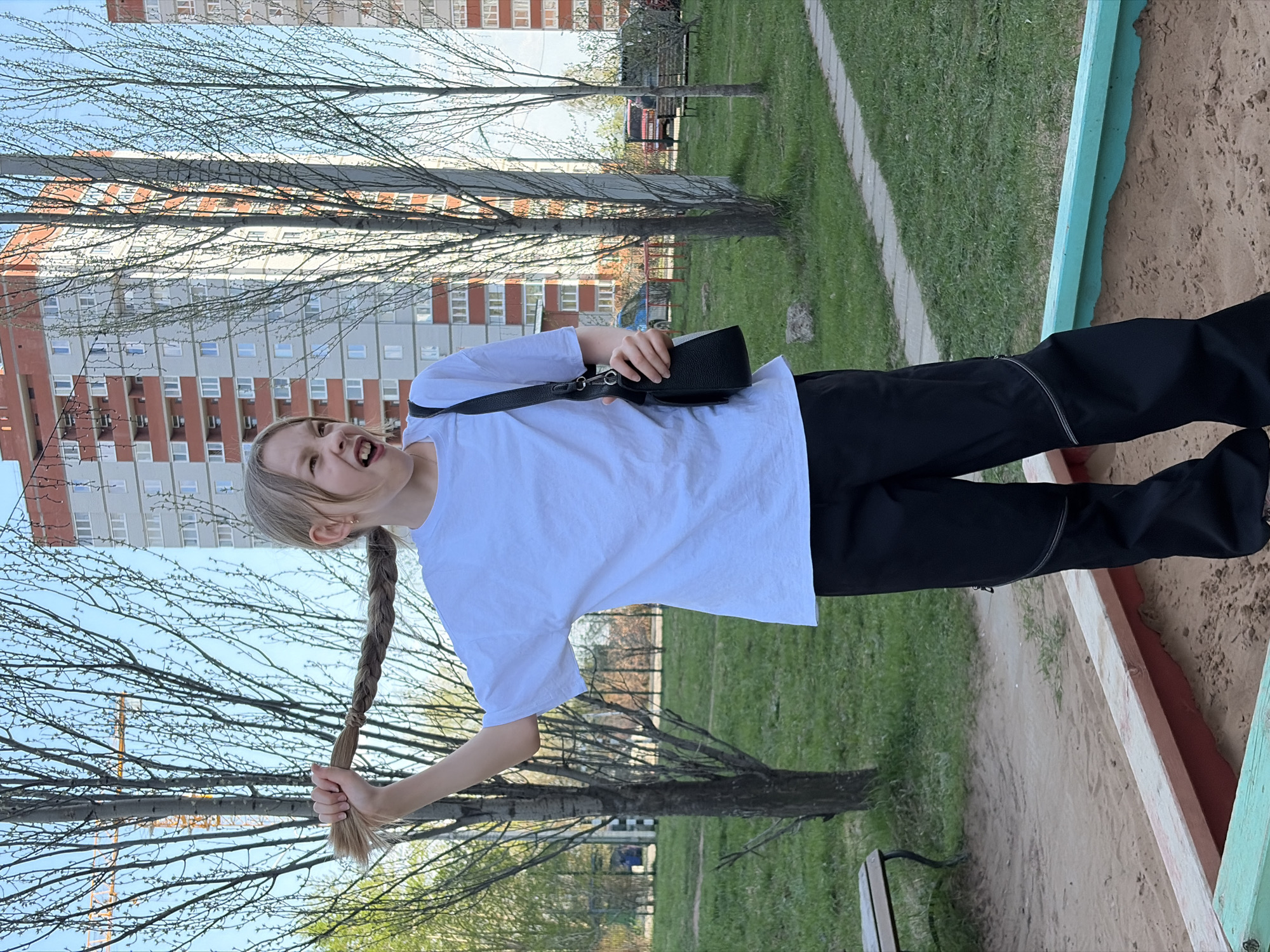 A girl with a long blonde braid stands near a sandbox in a park. She is wearing a white t-shirt, black pants, and a black shoulder bag. She is holding the end of her braid and making a silly face. In the background, there is a large apartment building, typical of the urban architecture in Izhevsk, Russia. The sky is blue, and there are trees with budding leaves.