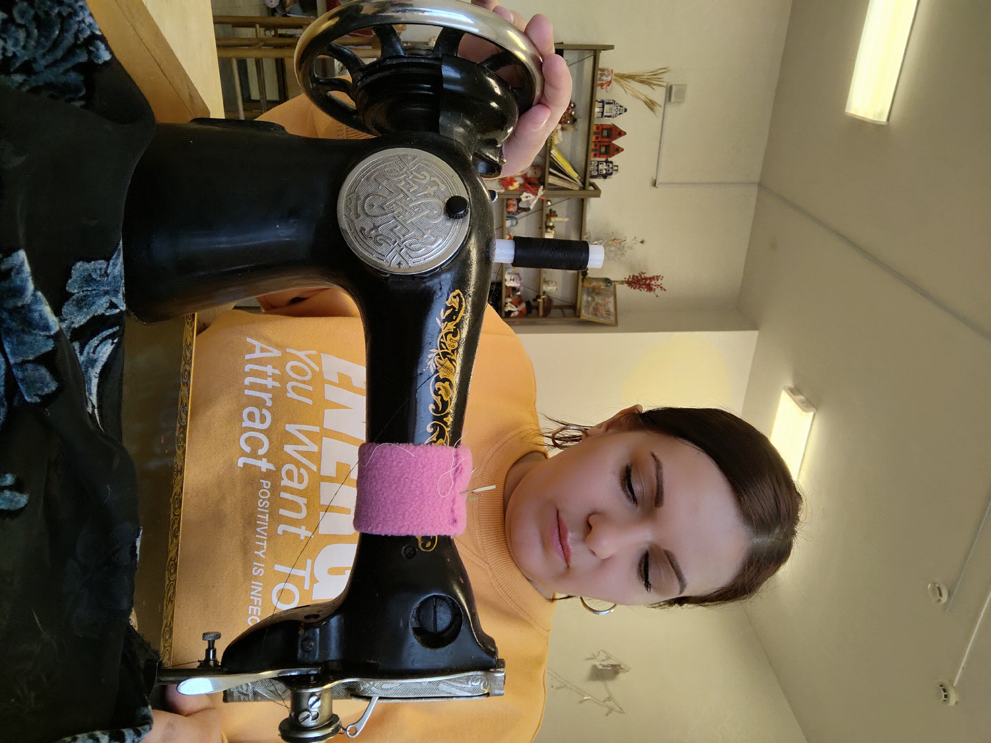 A woman is depicted indoors, engrossed in operating a vintage-style black sewing machine. She has dark hair, styled neatly, and her eyes are cast downwards, showing deep concentration on her task. She is wearing an orange or yellow long-sleeved shirt with partial text visible.
The black sewing machine, adorned with gold decorative accents and a silver handwheel, is the central object. A pink fabric band with needles is wrapped around the arm of the machine, serving as a pincushion. A black spool of thread is visible on a spindle towards the back of the machine's head. Dark fabric with a blue floral pattern is positioned under the needle, indicating that she is actively sewing. Her right hand is on the machine's handwheel, suggesting she is either turning it or has just made an adjustment.
In the background, behind the woman and the sewing machine, there is a light-colored wall with a shelf holding various decorative items, including small, colorful house models and bundles of dried straw or wheat. A bright rectangular fluorescent light fixture is visible on the light-colored ceiling.
The overall setting appears to be a well-lit indoor space, likely a home or a crafting area in Markovskii, Russia. The woman's expression conveys focus and diligence.
Visible text on her shirt reads: "You Want To Attract POSITIVITY IS INFECTIOUS".