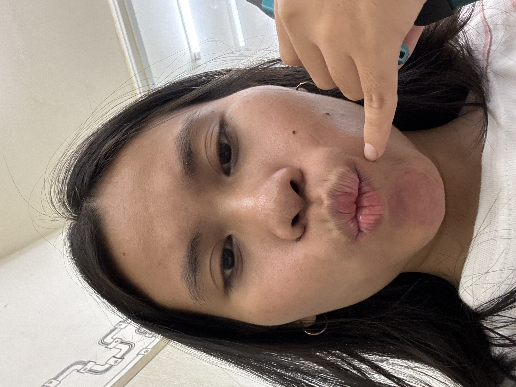 A close-up selfie of a young woman with dark hair and a light complexion is shown. She is making a duck face, with her lips pursed. Her right index finger is touching her left cheek, in a gesture that can resemble a phone call. She is wearing a white t-shirt with thin pink stripes. The background appears to be an indoor environment, possibly a hallway, with a white wall featuring a decorative patterned line and ceiling lights. The image was taken in East Jakarta, Indonesia.