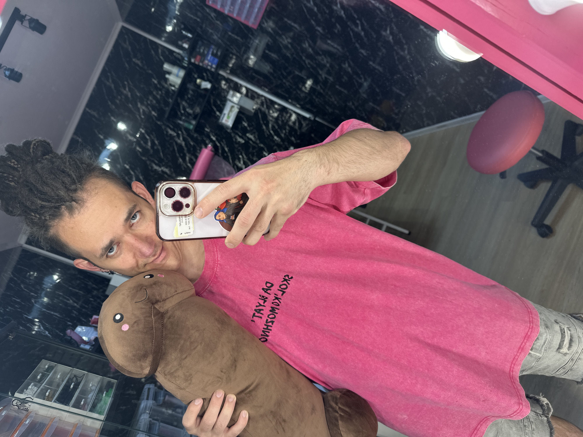 A person with dreadlocks is taking a selfie in a room with a glossy black marble-patterned wall and a bright pink trim. They are wearing a pink t-shirt and holding a brown, phallus-shaped plush toy with a smiling face. The person is using a smartphone with a tan case and stickers on it to capture the image. The room contains shelving with various items, and a partially visible chair with a pink seat. Based on the t-shirt text, the location could potentially be the city of Saratov, Russia.
