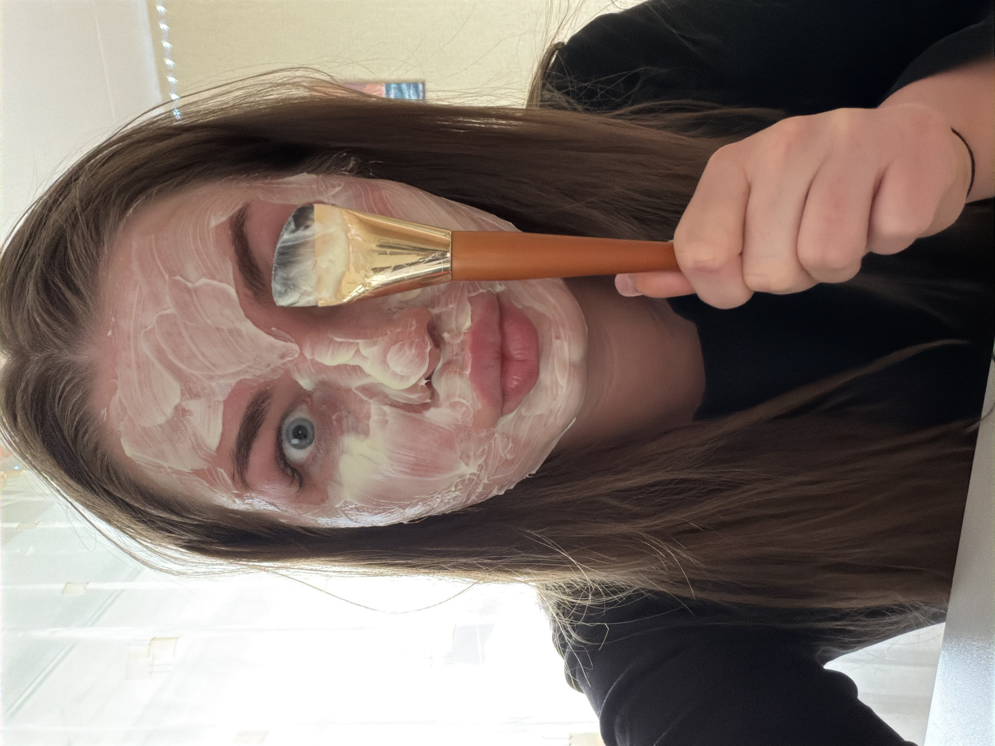 A young woman with long brown hair is applying a white face mask with a flat makeup brush. The mask covers most of her face except for her lips. She is wearing a black shirt. The background appears to be a room with a window. There is no information to suggest the photo was taken in the city of Chaikovsky, Russia.
