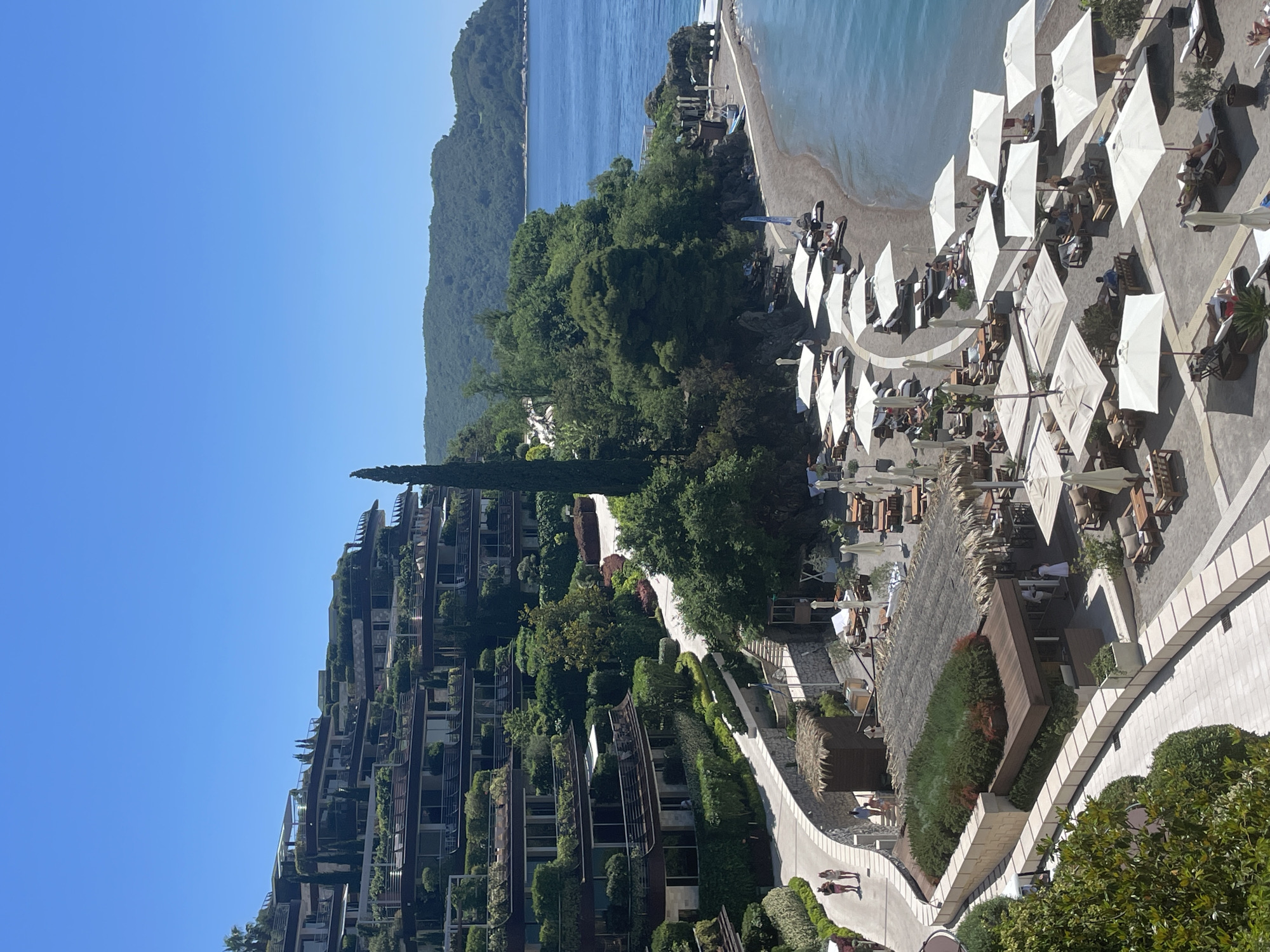 This image shows an elevated view of a coastal resort in the city of Budva, Montenegro. The scene includes a beach with rows of white umbrellas and sun loungers occupied by people relaxing. The beach meets the turquoise waters of the Adriatic Sea. A green, mountainous landscape stretches along the coastline in the background. Terraced buildings with balconies adorned with greenery rise up the slope, cascading towards the beach. A pathway meanders through the resort, lined with manicured hedges and landscaping. People can be seen strolling along the pathway. The scene is bathed in bright sunlight, indicating a clear, sunny day.